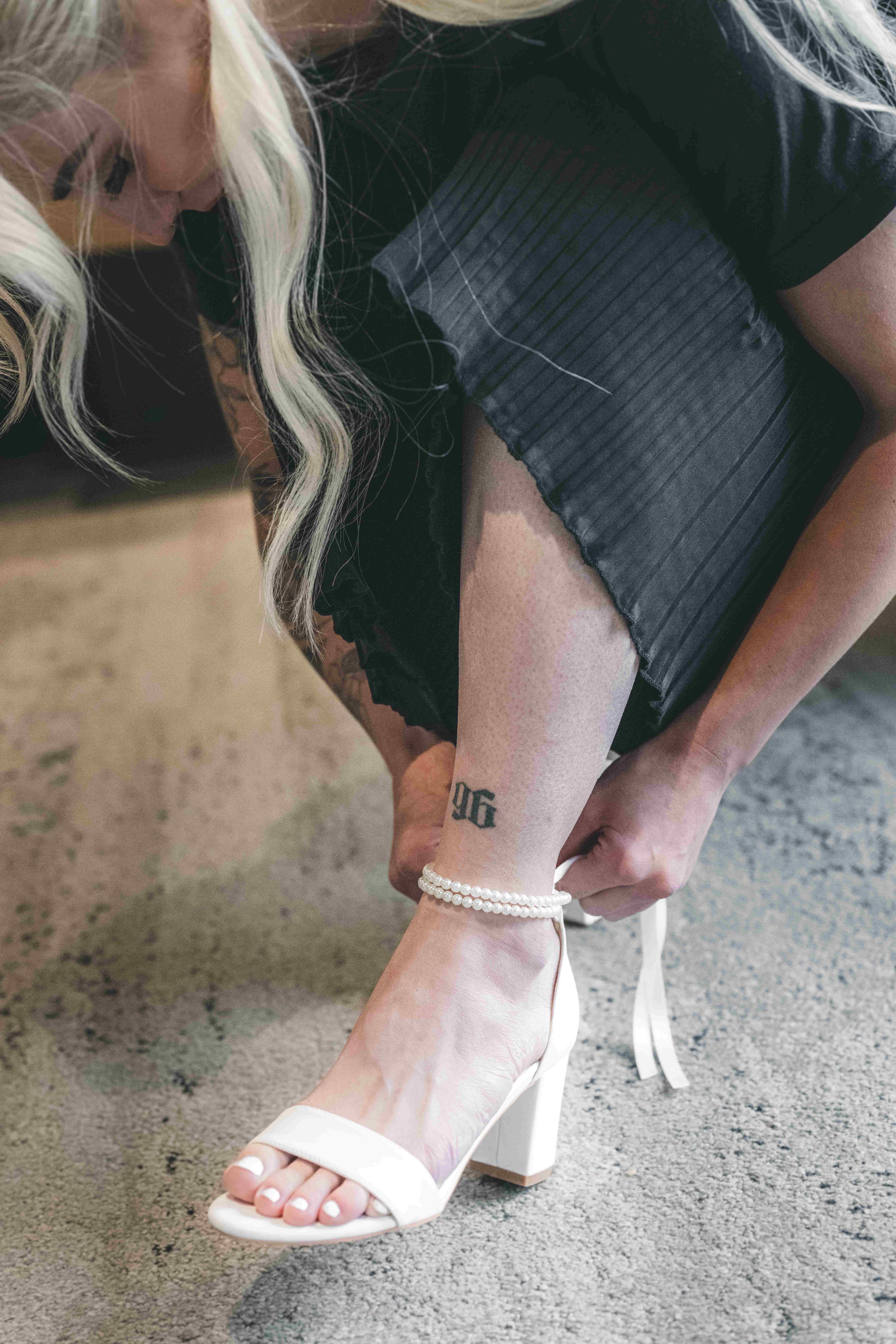 Die Braut beugt sich nach vorne, um das feine Perlenriemchen ihres weißen Brautschuhs zu schließen, während ihr Tattoo am Bein zu sehen ist.