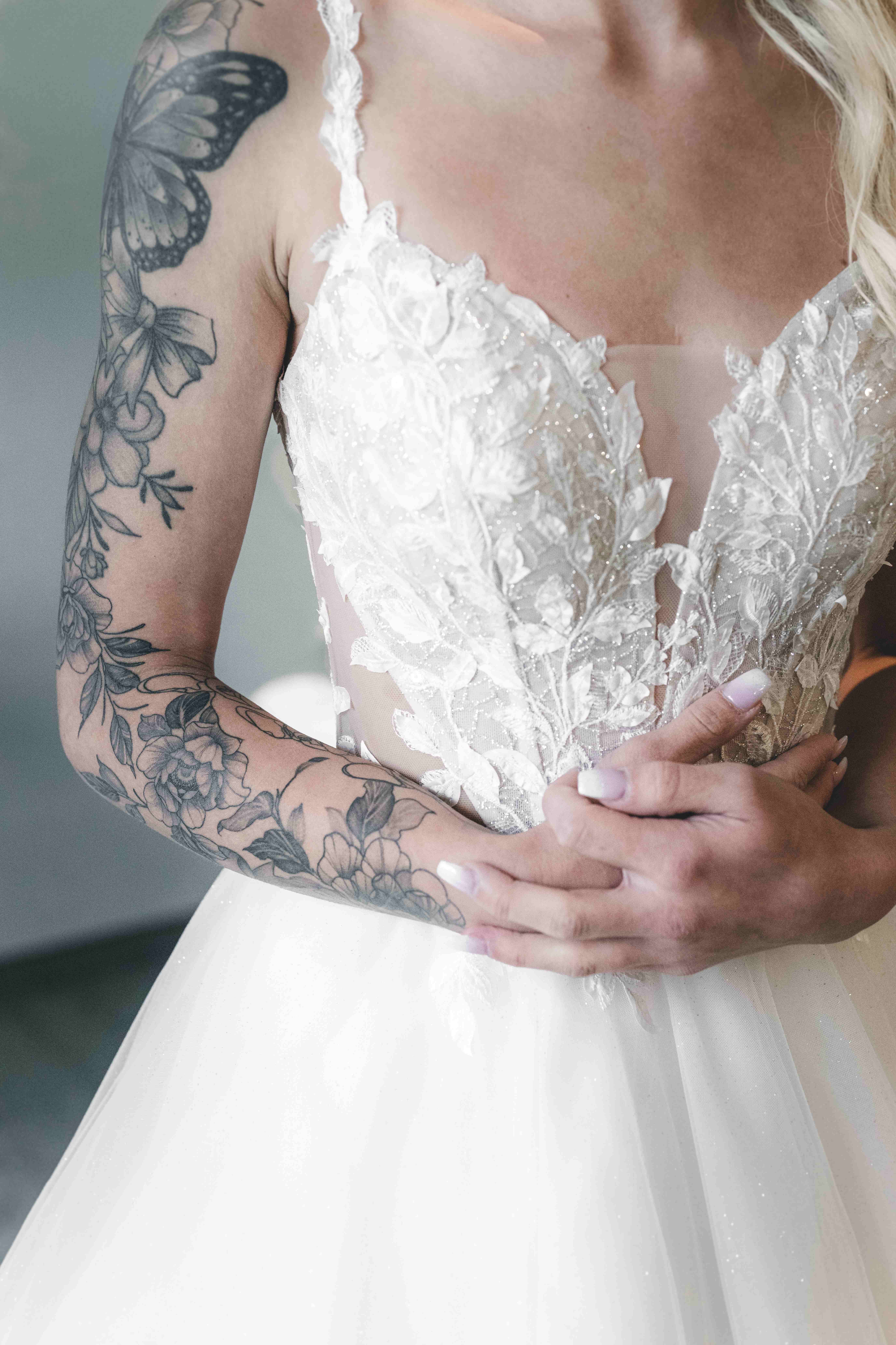 Nahaufnahme der filigranen, floralen Spitze und des tiefen Ausschnitts am Brautkleid, während die Braut ihre Hände elegant verschränkt hält.