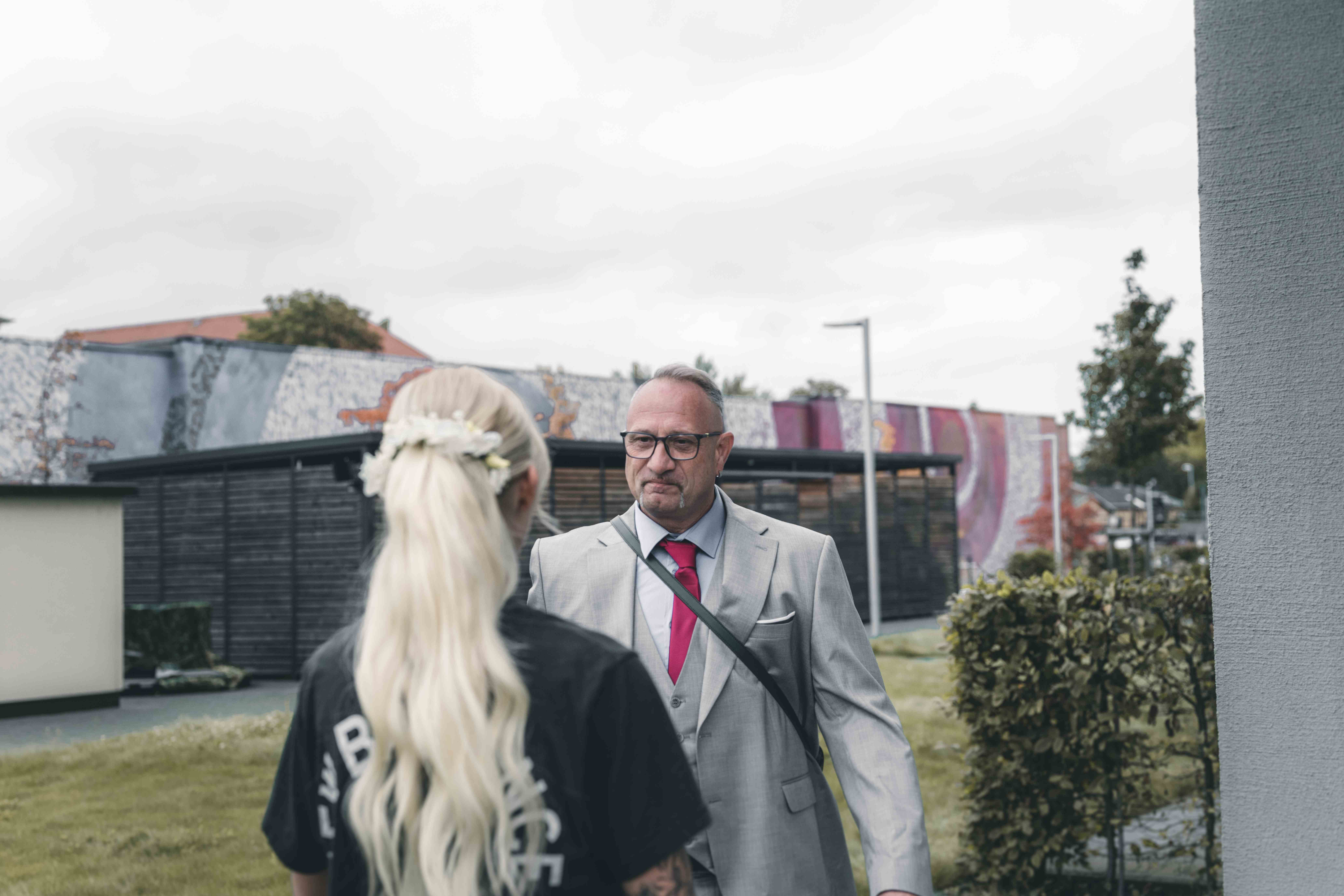 Ein sichtlich gerührter Brautvater im grauen Anzug blickt seine Tochter (von hinten zu sehen) beim ersten Aufeinandertreffen vor der Trauung voller Stolz an.