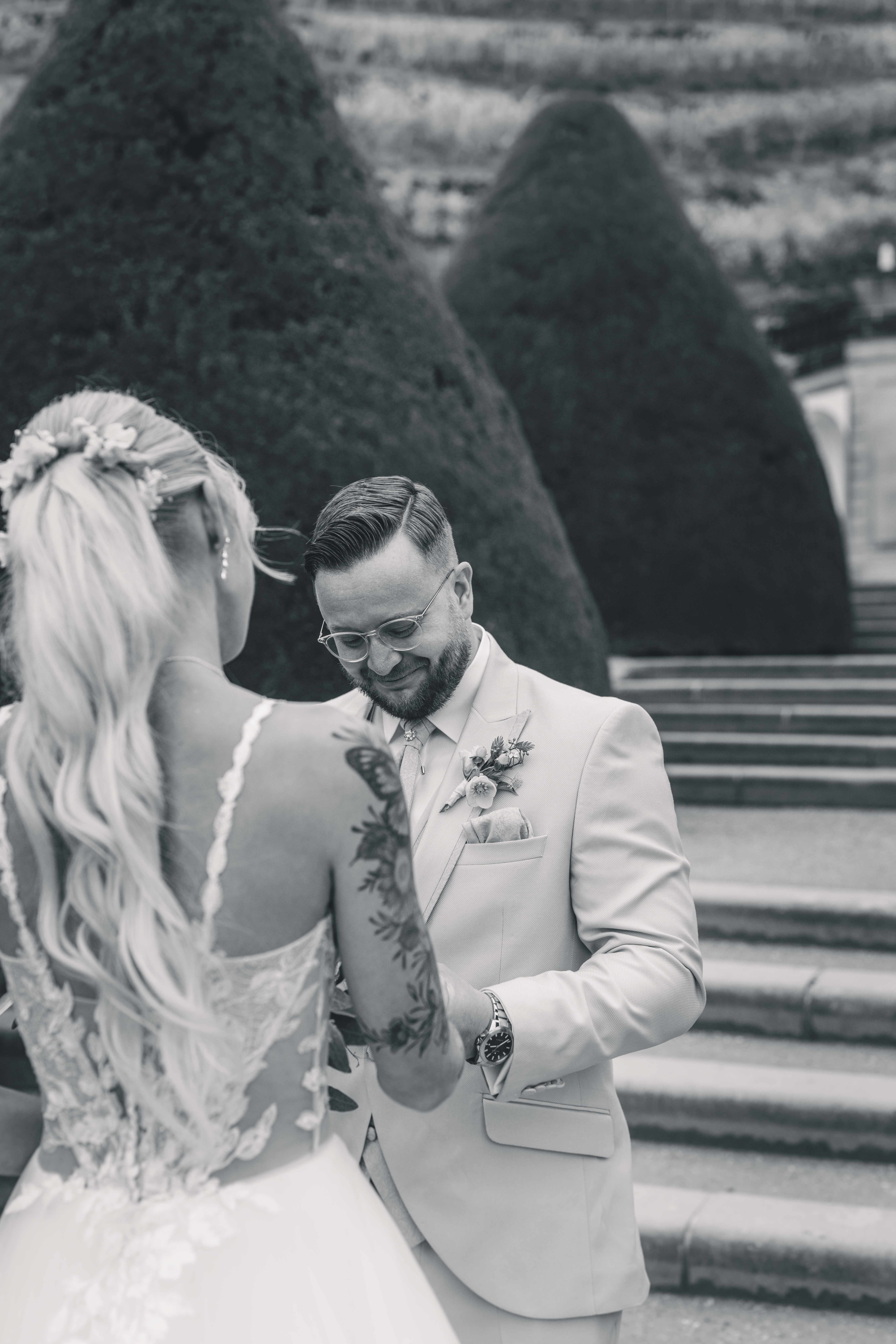 Schwarz-Weiß-Aufnahme eines Bräutigams mit Brille, der beim First Look im Schlossgarten gerührt zu Boden blickt und lächelt.