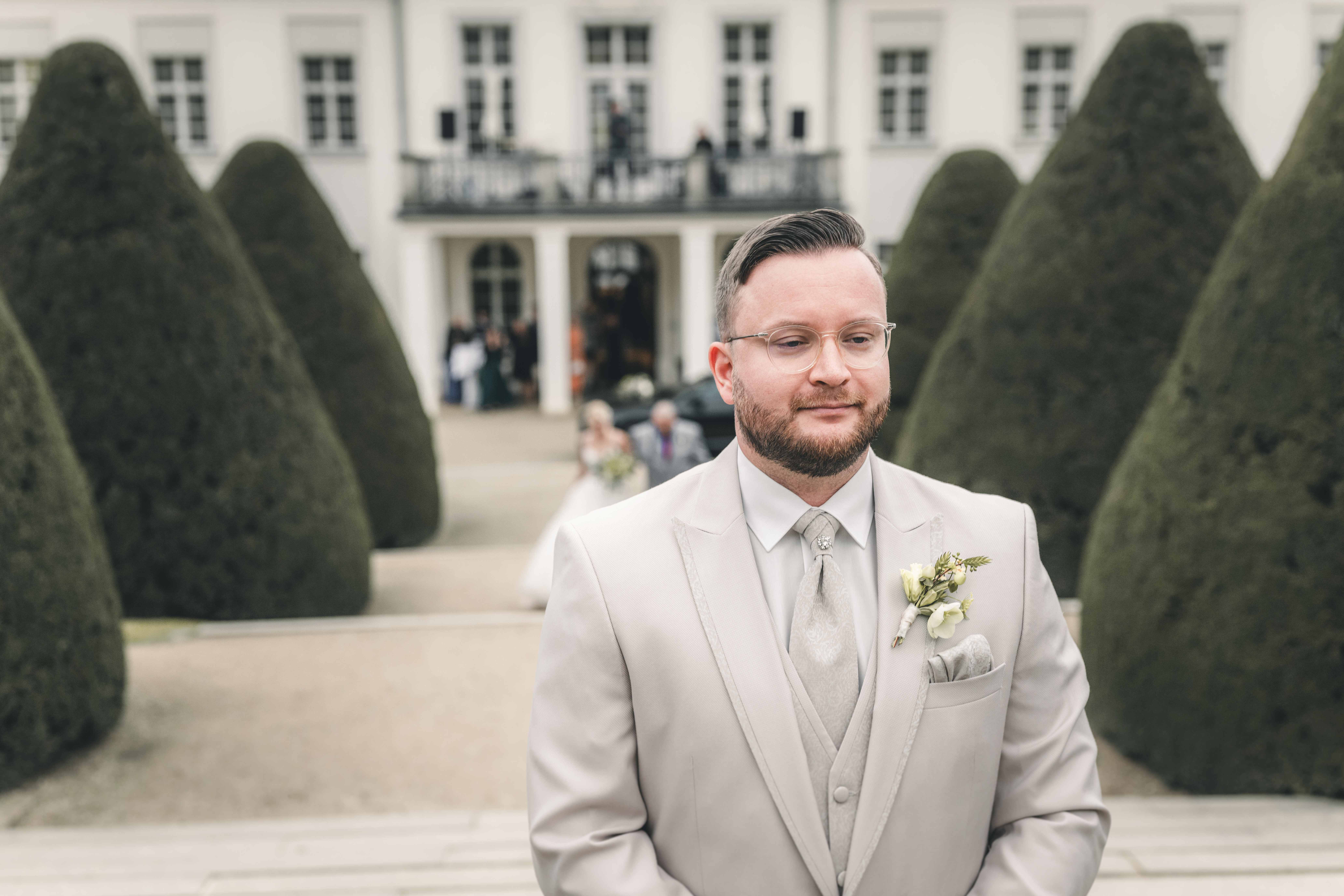 Der Bräutigam wartet im hellen Anzug im Barockgarten von Schloss Wackerbarth, während die Braut im Hintergrund von ihrem Vater zu ihm geführt wird.
