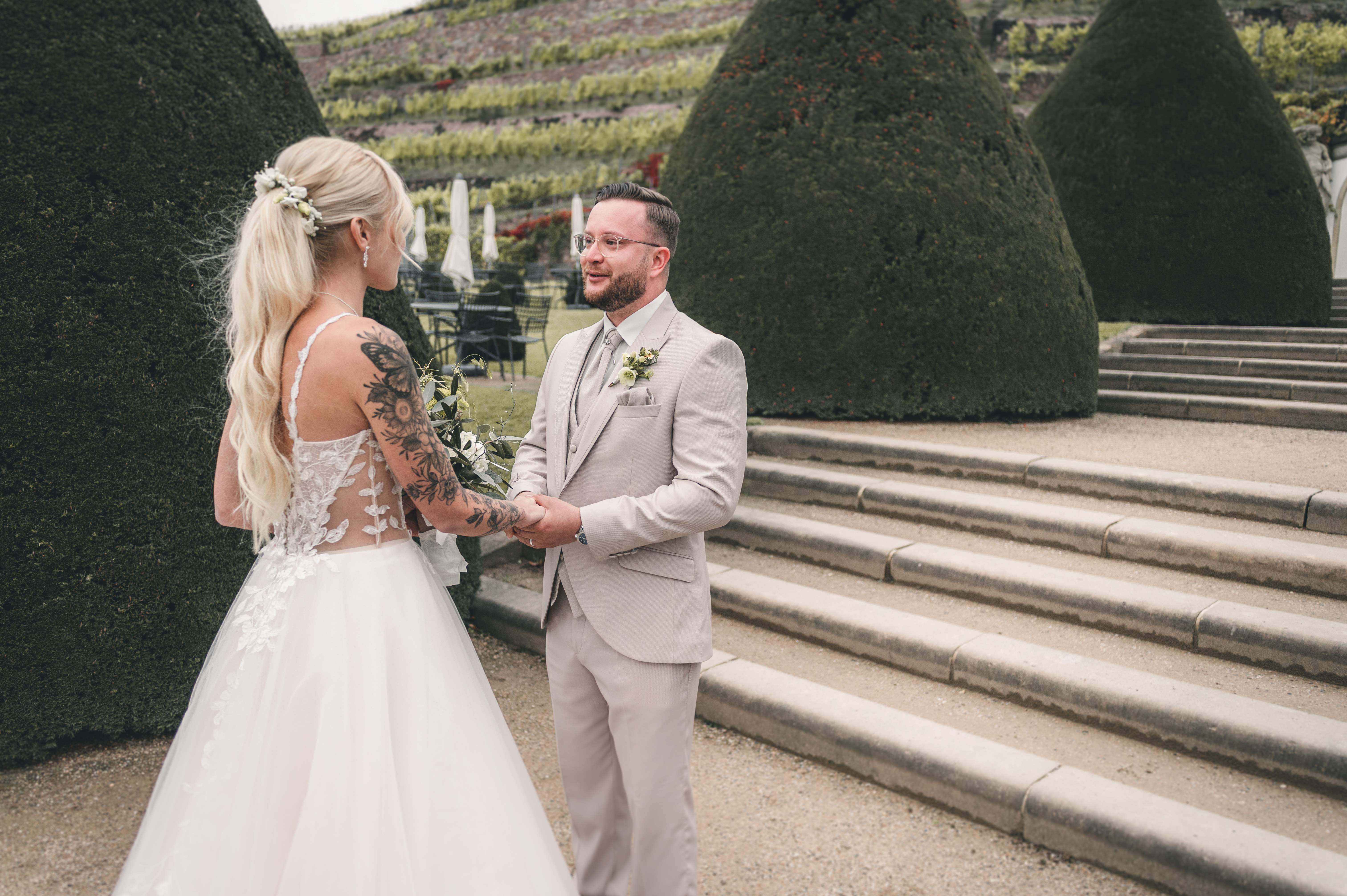 Das Brautpaar hält im Schlossgarten von Schloss Wackerbarth vor den terrassenförmigen Weinbergen und großen Buchsbaumhecken Händchen beim First Look.