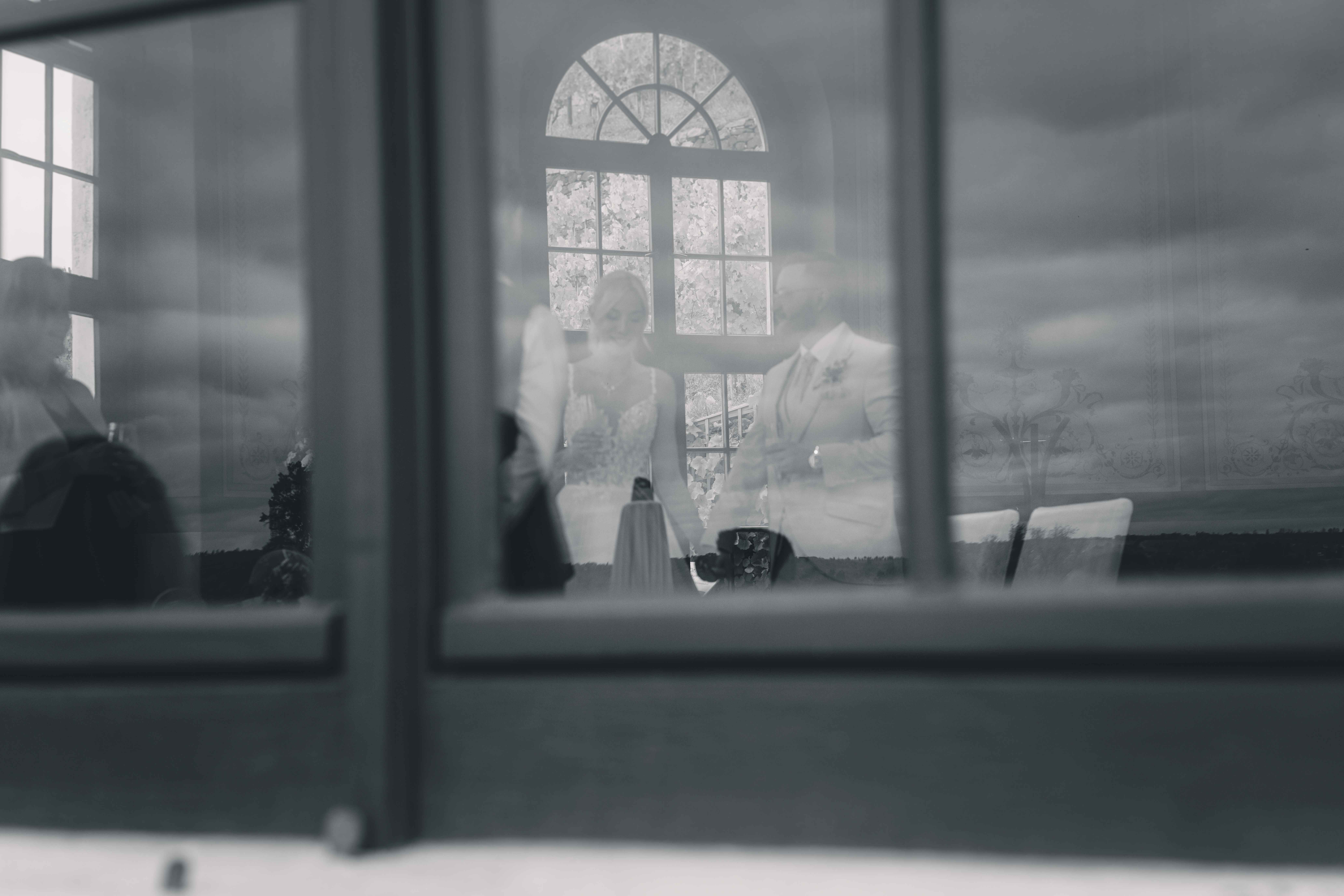 Eine kunstvolle Schwarz-Weiß-Fotografie, die das Brautpaar als sanfte Spiegelung in den Fensterscheiben des Schlosses zeigt, während im Hintergrund die Weinberge zu sehen sind.