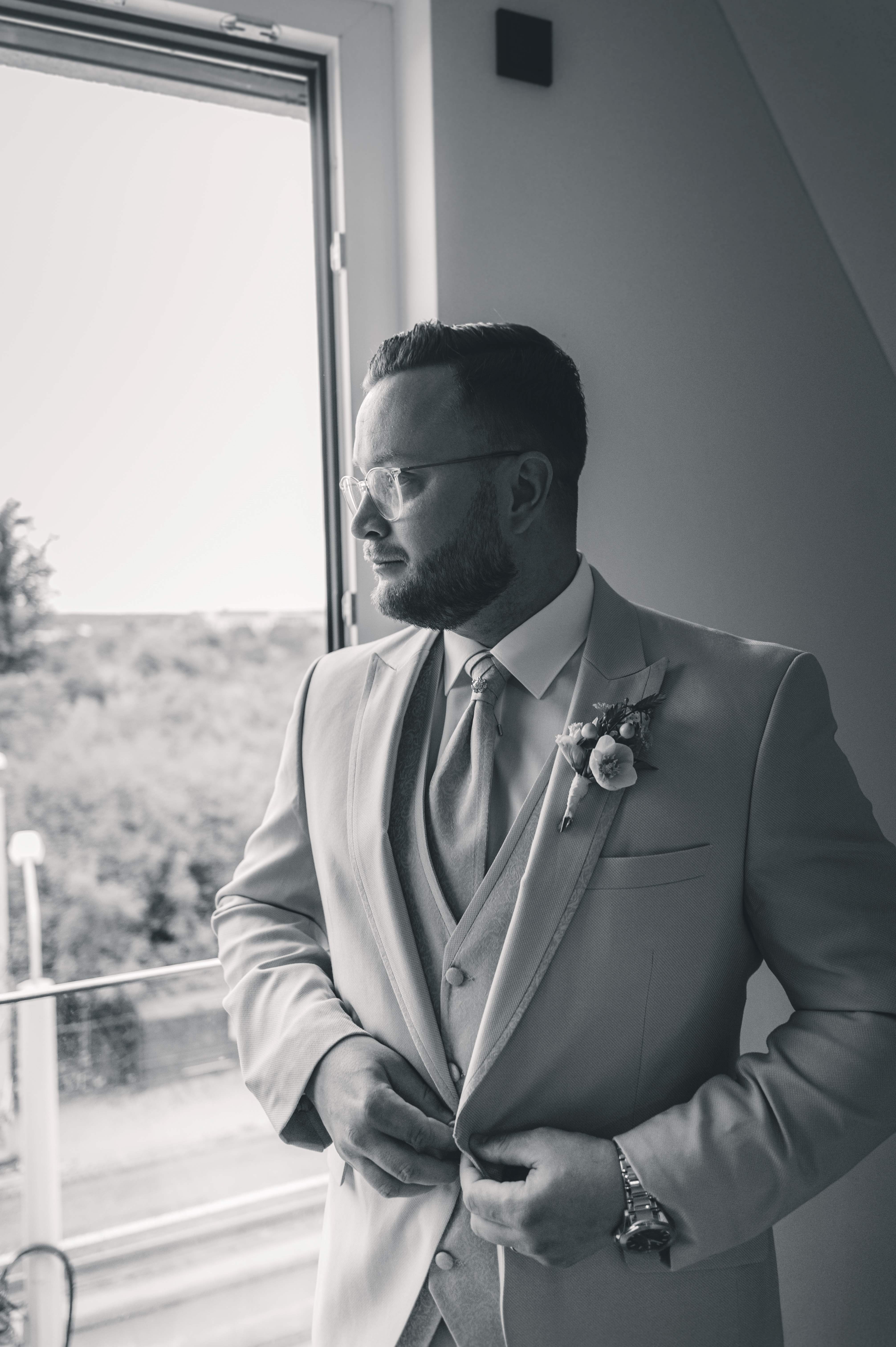 Eine stimmungsvolle Schwarz-Weiß-Aufnahme des Bräutigams im Profil, der in seinem Hochzeitsanzug nachdenklich und voller Vorfreude aus dem Fenster blickt.