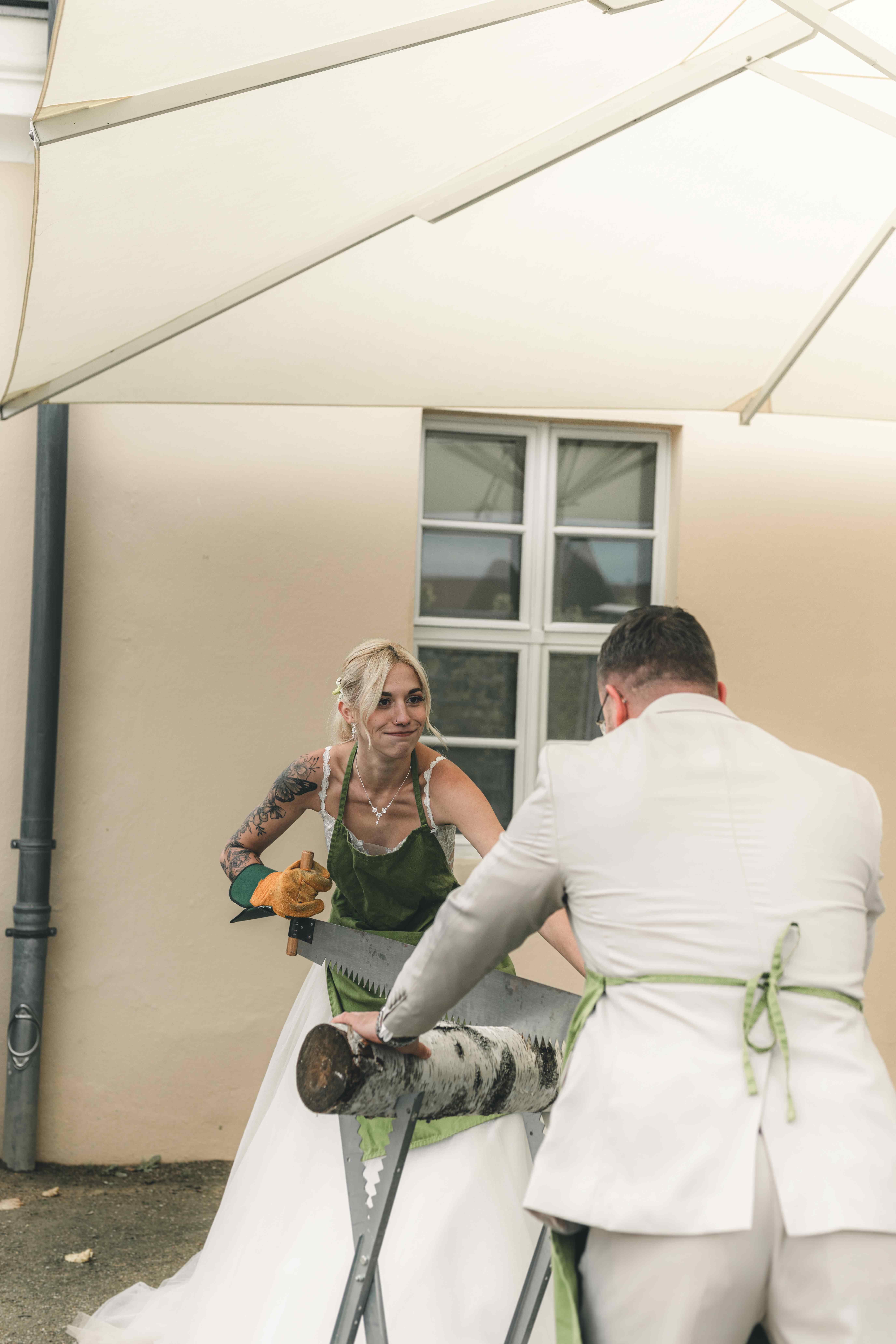 Das Brautpaar in Hochzeitskleidung und grünen Schürzen sägt gemeinsam voller Elan mit einer großen Zugsäge einen Birkenstamm im Außenbereich von Schloss Wackerbarth durch.
