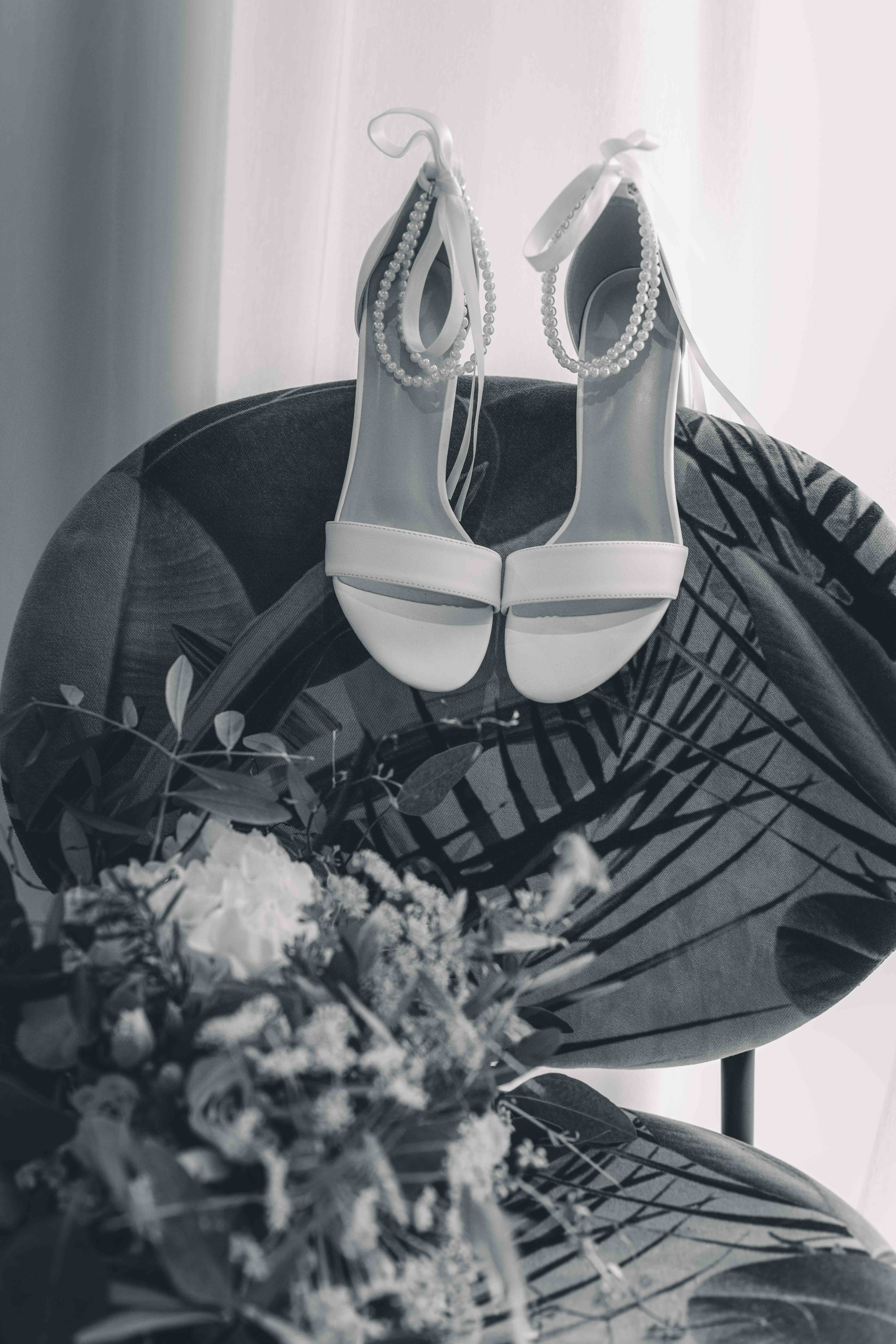 Eine stimmungsvolle Schwarz-Weiß-Aufnahme von zwei eleganten weißen Brautschuhen mit Perlenkette und Satinschleife, die an einer Stuhllehne hängen.
