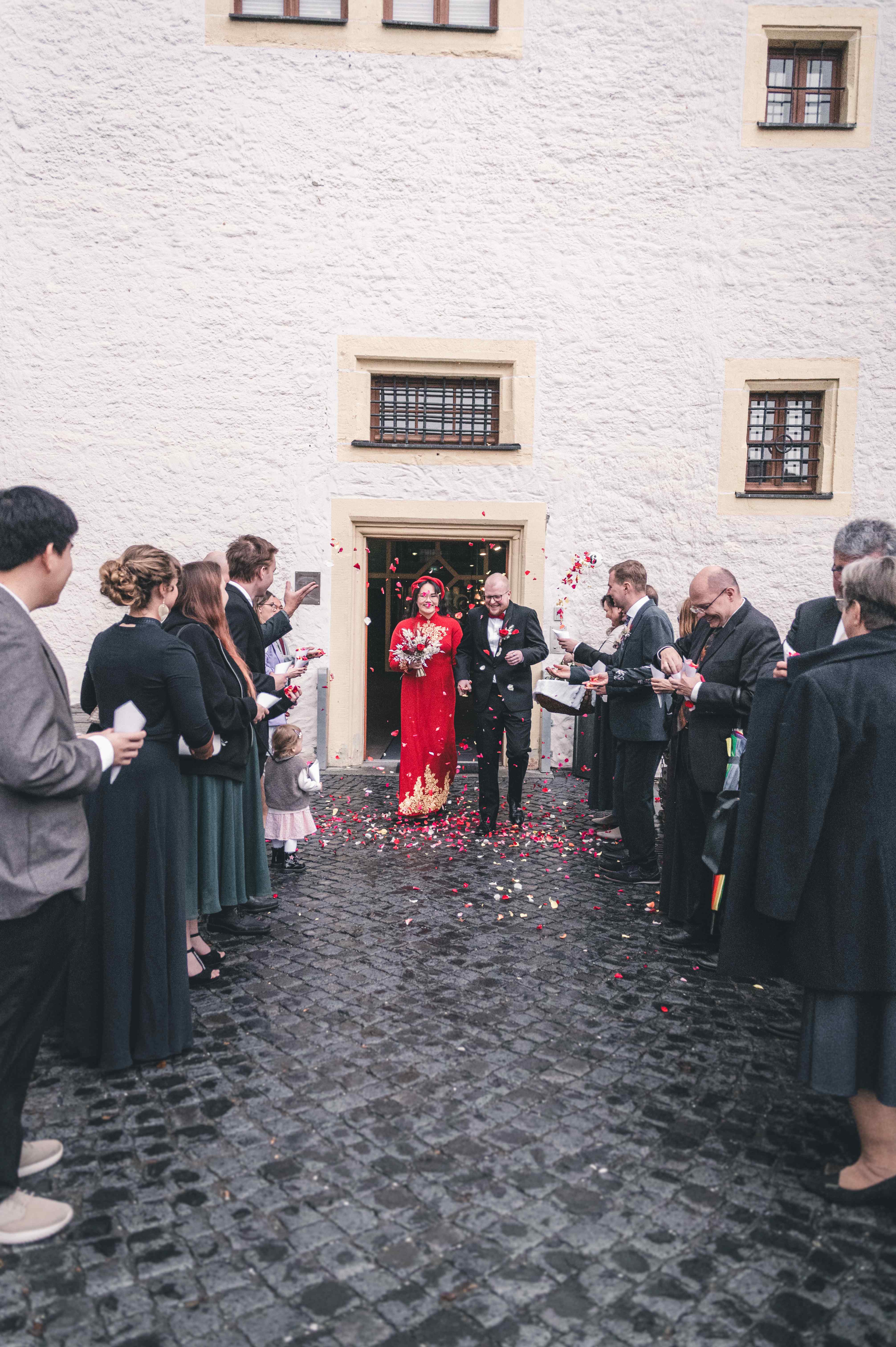 Das frischgebackene Ehepaar schreitet Hand in Hand durch das Portal von Schloss Klaffenbach, während Gäste rote Blütenblätter werfen.