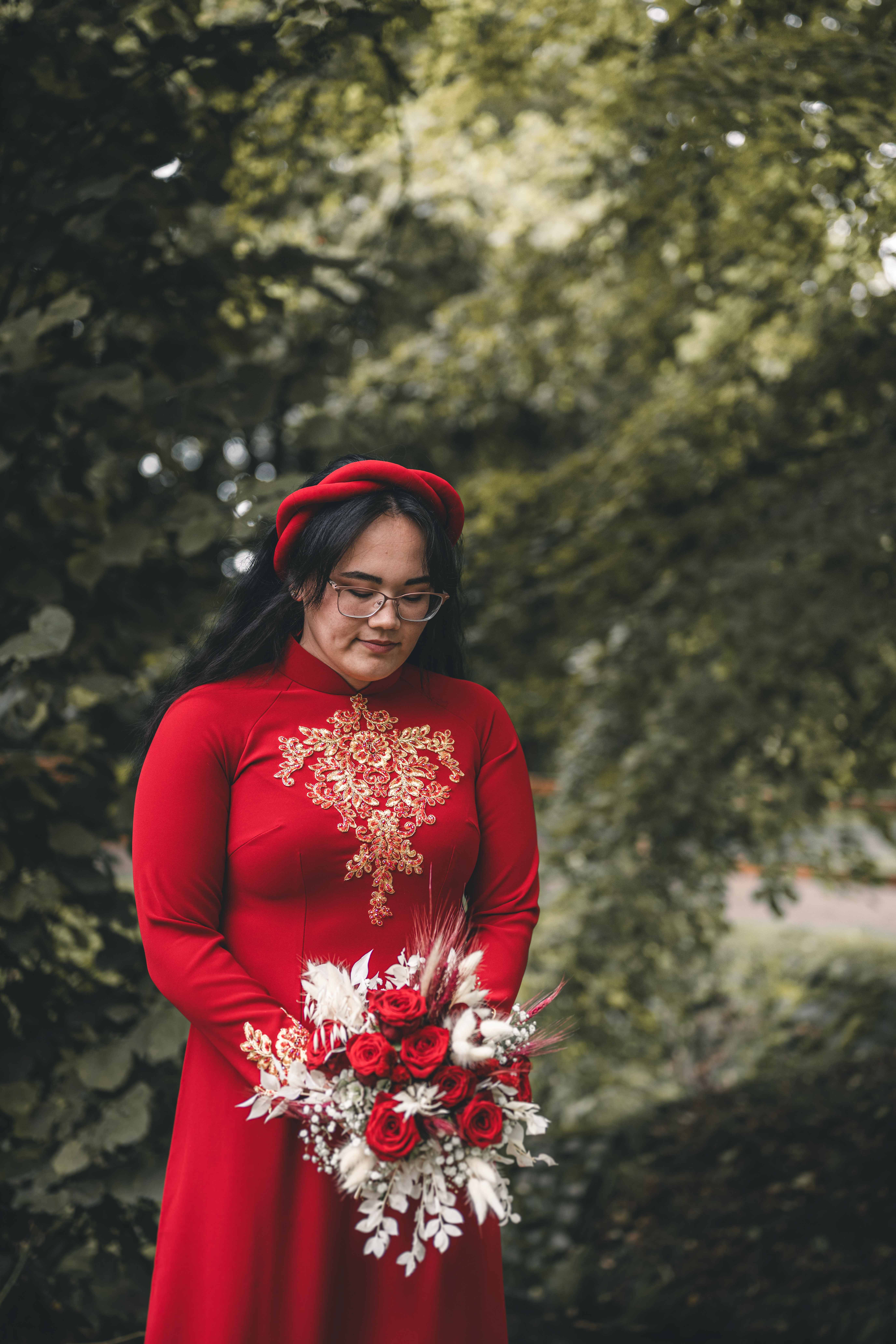 Ein elegantes Portrait der Braut, die im roten vietnamesischen Ao Dai mit goldenen Stickereien im Schlosspark Klaffenbach steht und behutsam auf ihren Strauß blickt.