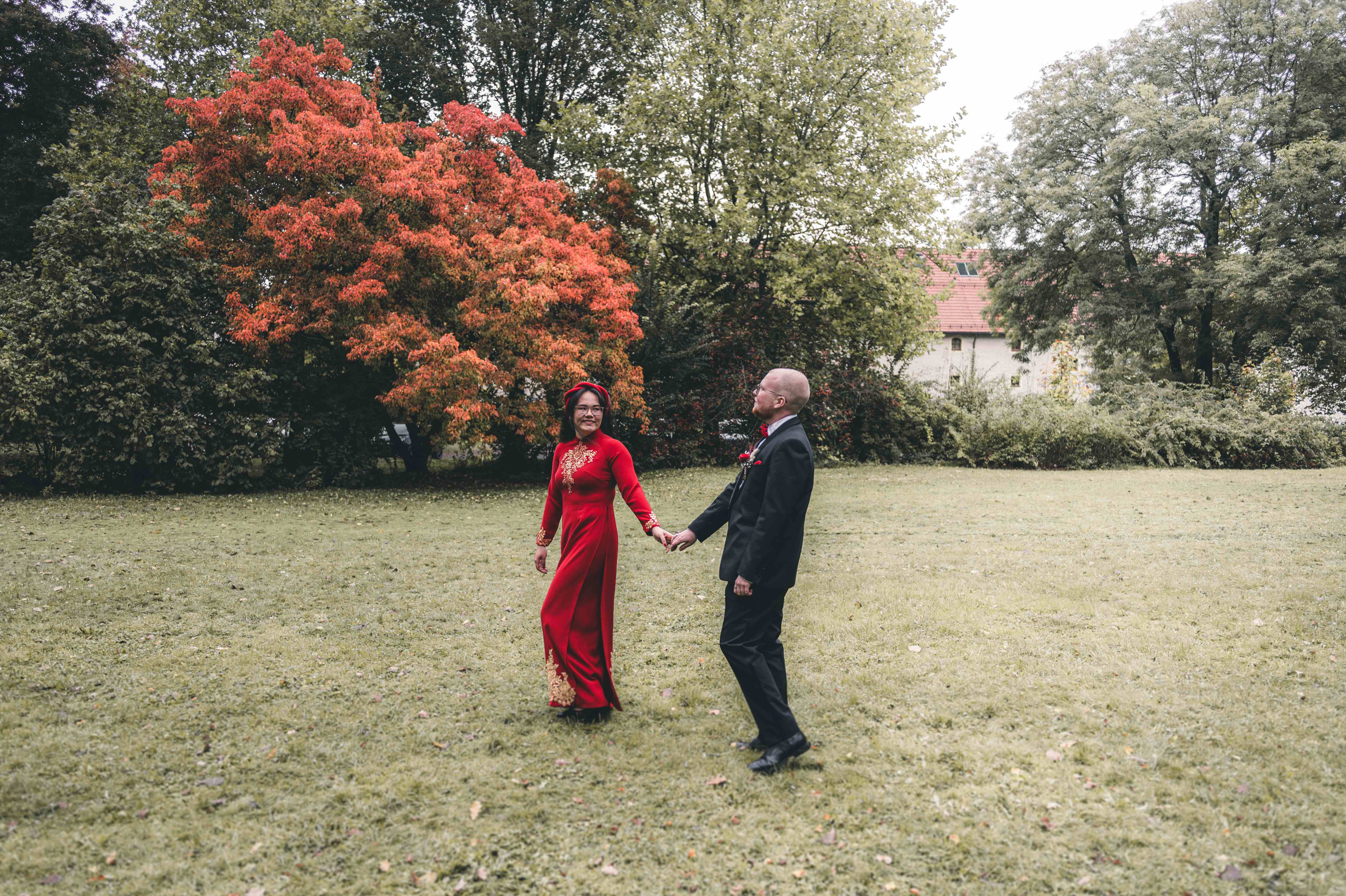 Das Brautpaar läuft Hand in Hand über eine grüne Wiese im Schlosspark Klaffenbach, im Hintergrund strahlt ein Baum in leuchtenden Herbstfarben.