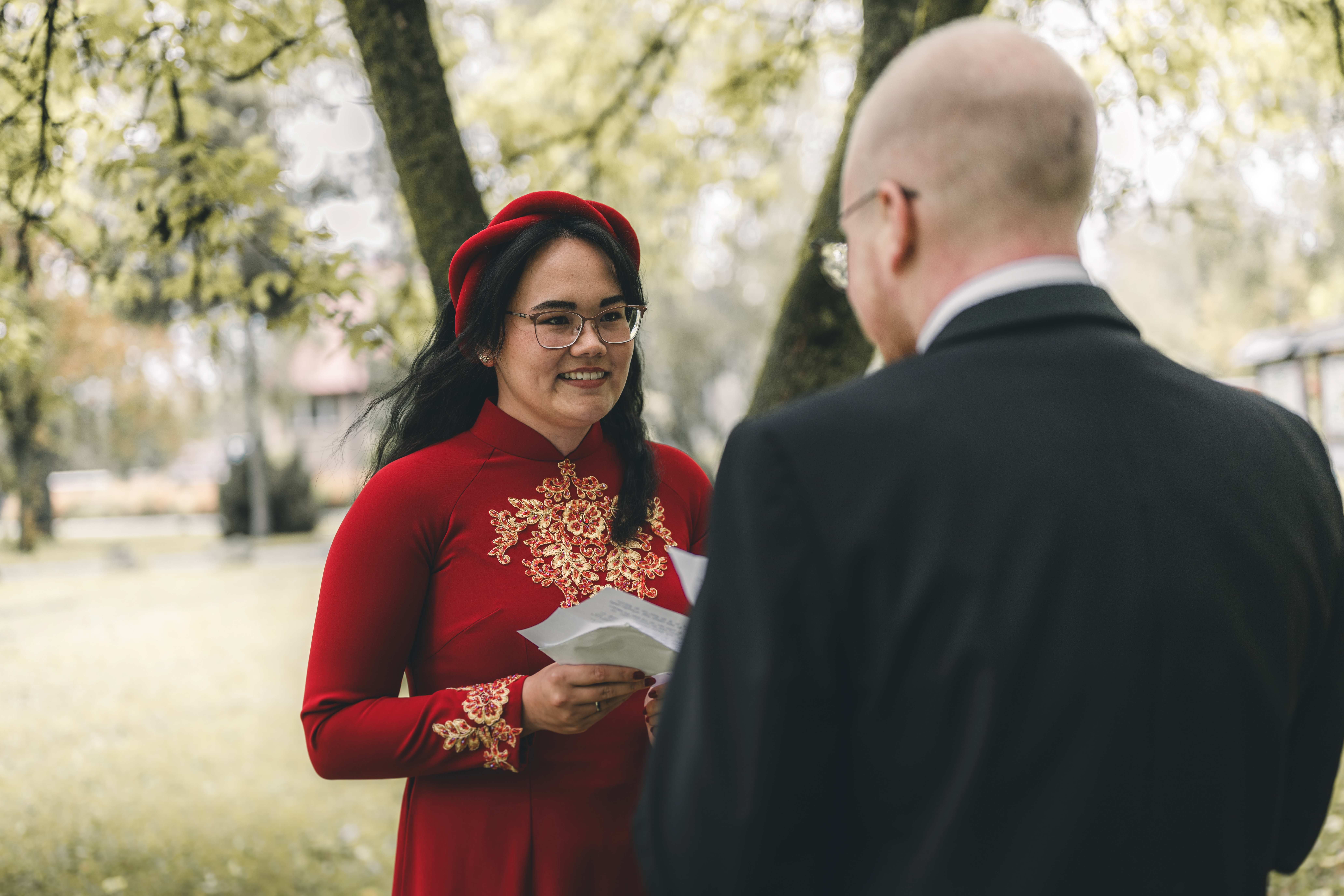 Die Braut im traditionellen roten vietnamesischen Kleid liest dem Bräutigam im Schlossgarten Klaffenbach bei Chemnitz lächelnd ihr Eheversprechen vor.