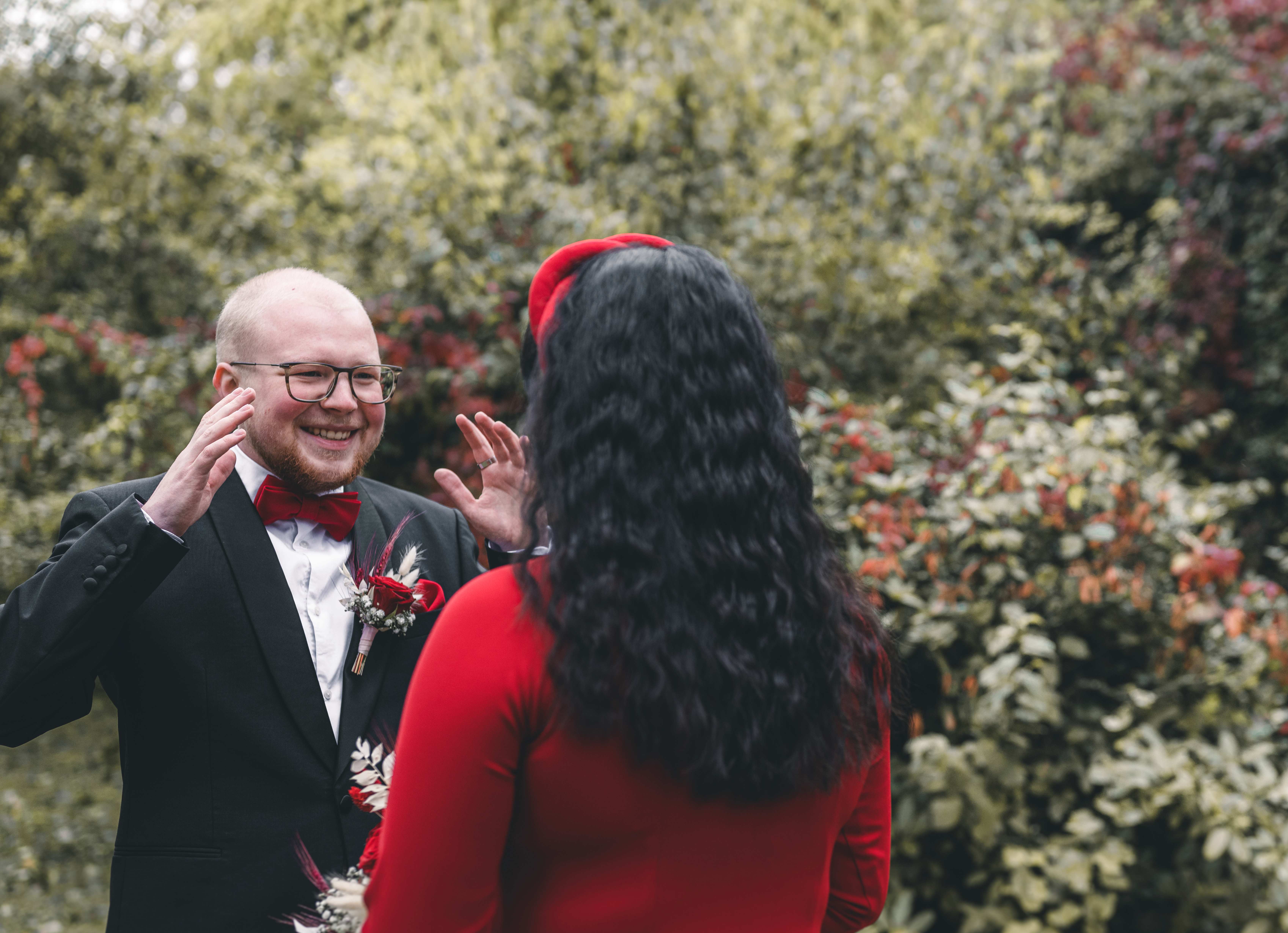 Der Bräutigam im schwarzen Anzug hebt beim First Look im Schlossgarten Klaffenbach fasziniert die Hände, als er seine Braut im roten Kleid zum ersten Mal sieht.