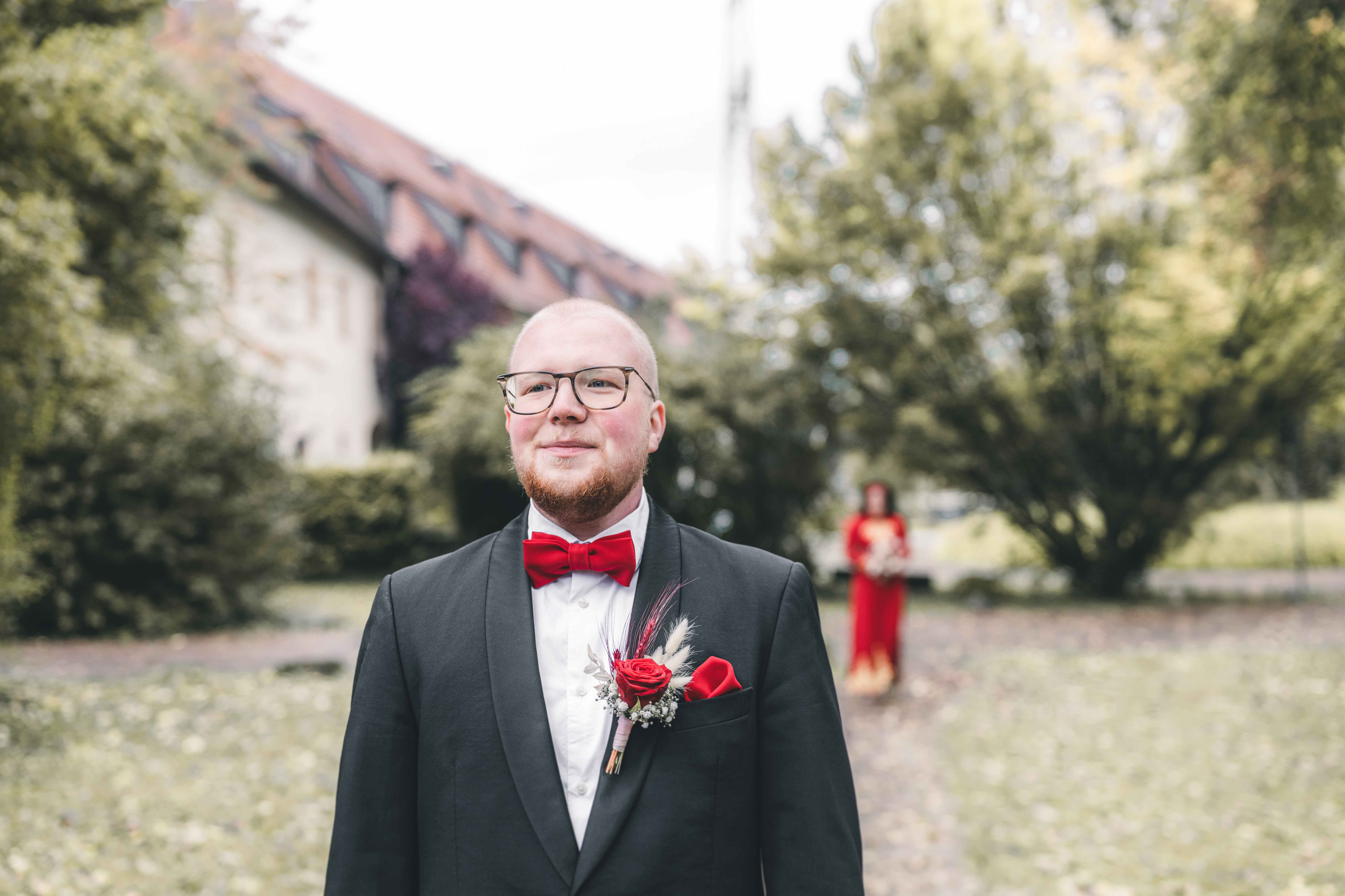 Der Bräutigam im Hochzeitsanzug wartet lächelnd im Park von Schloss Klaffenbach, während die Braut im roten Kleid im Hintergrund zu sehen ist.