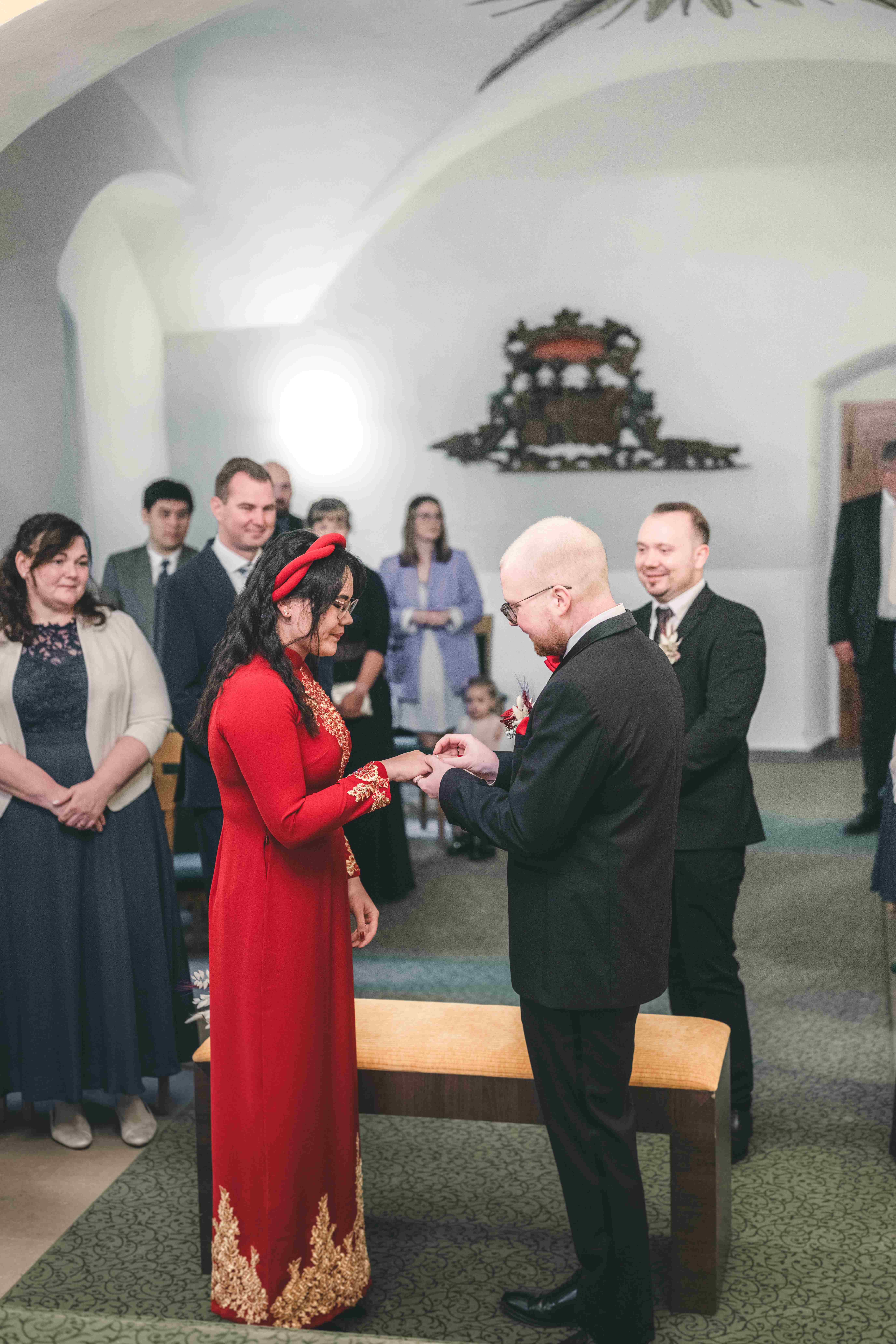Der Bräutigam steckt seiner Braut im roten traditionellen Kleid während der standesamtlichen Zeremonie in Sachsen lächelnd den Ehering an.