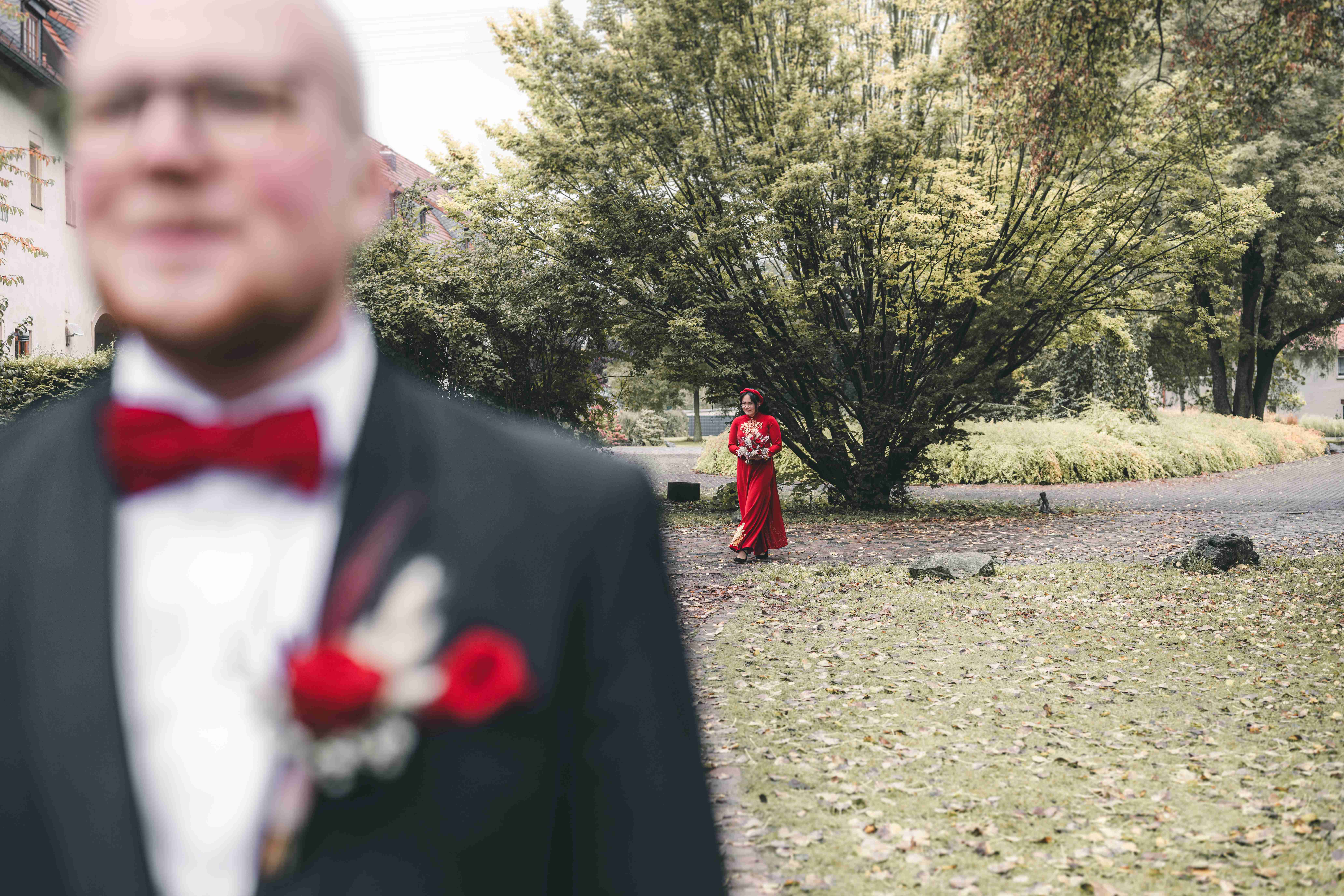 Die Braut im traditionellen roten vietnamesischen Hochzeitskleid (Ao Dai) läuft mit ihrem Strauß durch den Schlossgarten, im Vordergrund wartet der Bräutigam unscharf.