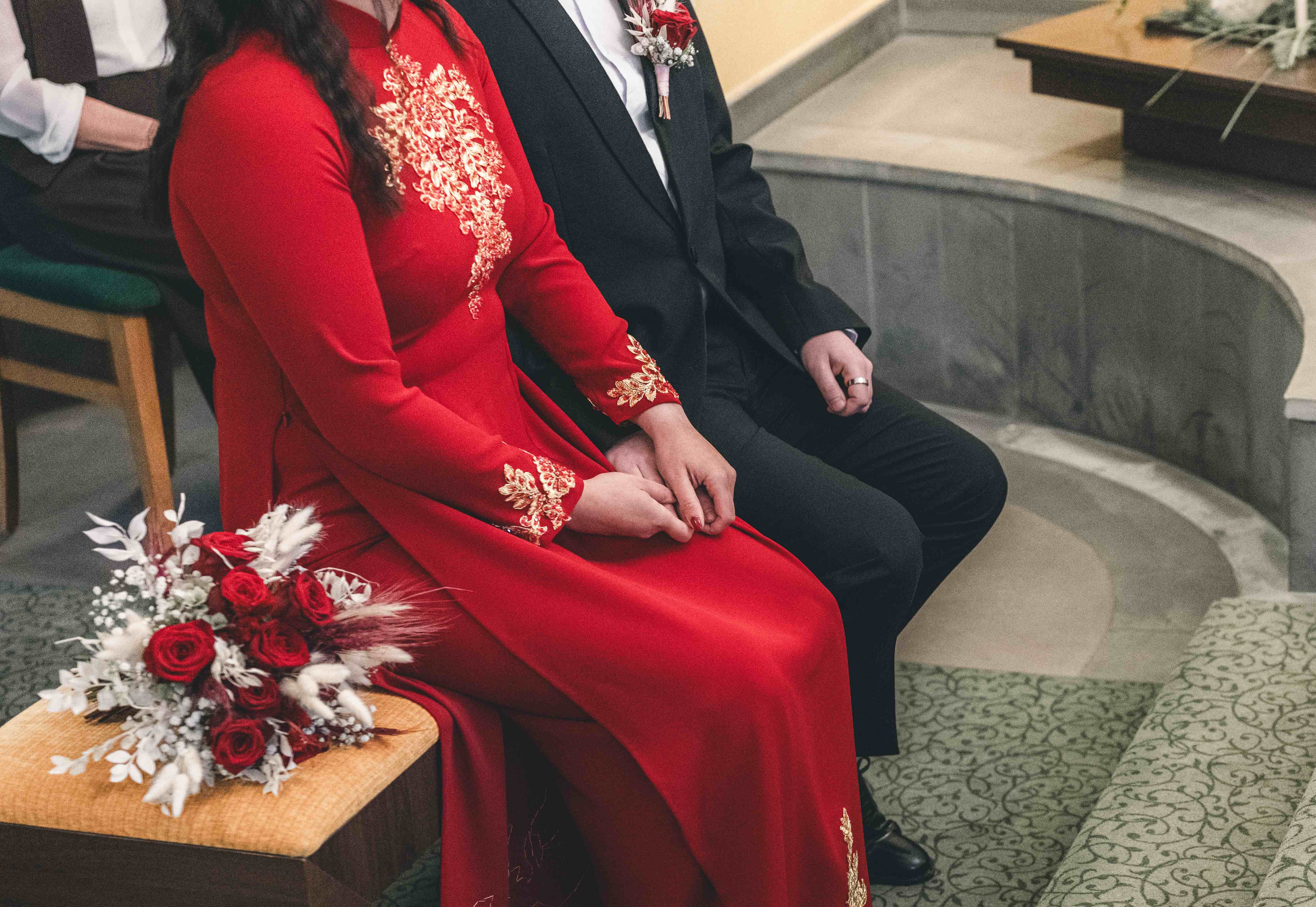 Nahaufnahme des Brautpaares auf den gelben Stühlen im Standesamt, der rot-weiße Brautstrauß liegt elegant auf einem Hocker im Vordergrund.
