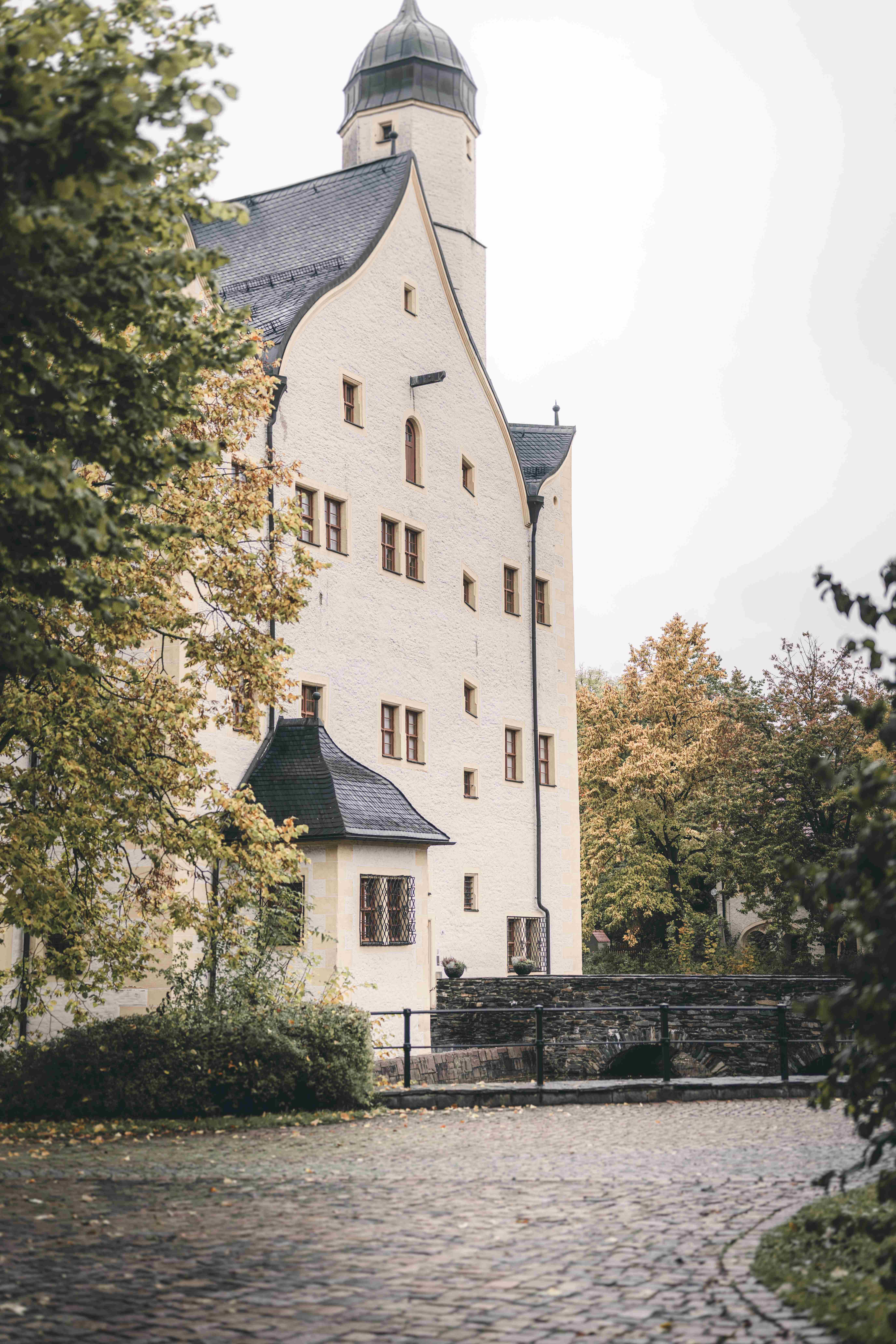 Außenansicht des weißen Wasserschlosses Klaffenbach bei Chemnitz, malerisch eingerahmt von herbstlichen Bäumen im Schlossgarten.