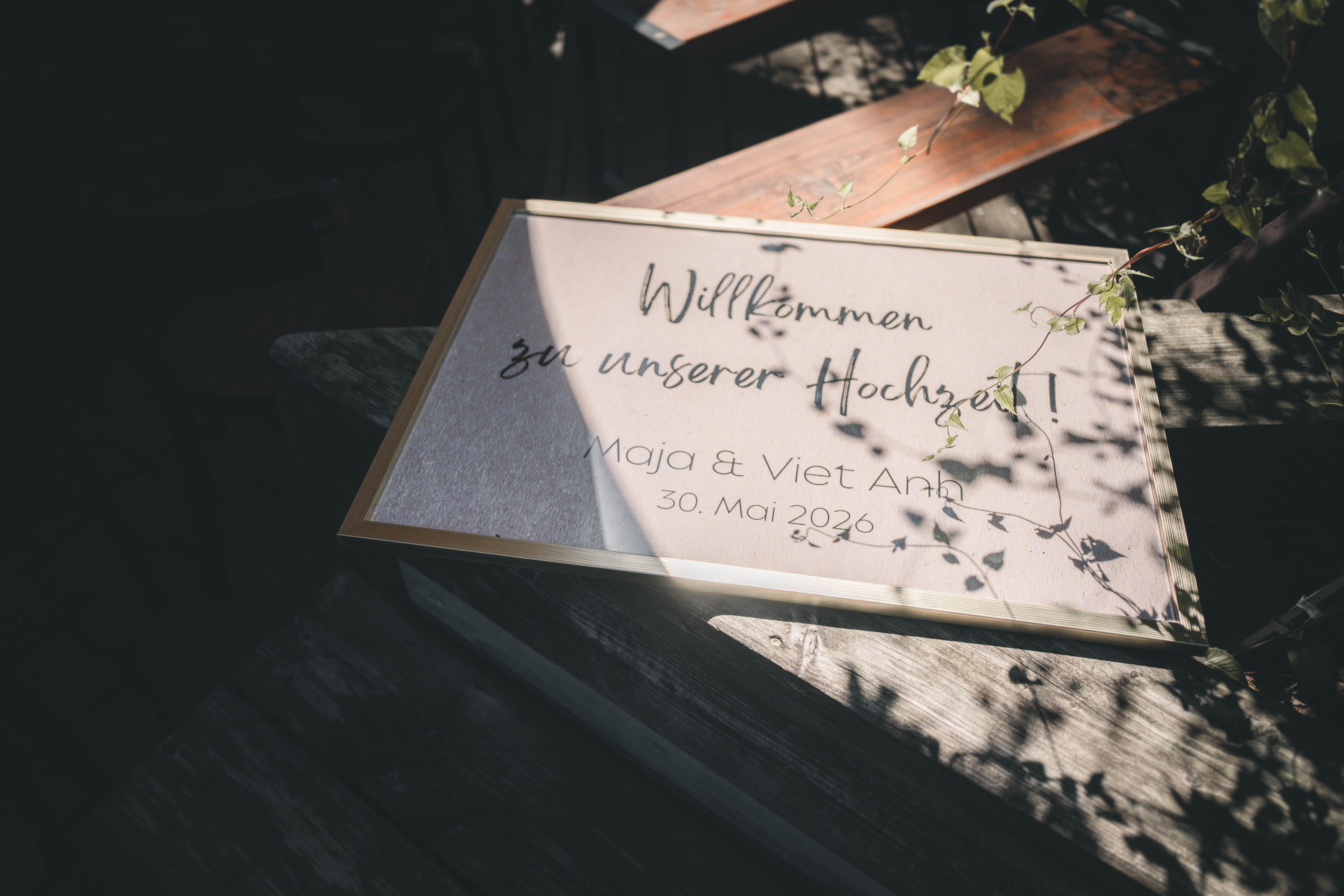 Das edle, selbstgestaltete Willkommensschild „Willkommen zu unserer Hochzeit! Maja & Viet Anh – 30. Mai 2026“ liegt im Spiel von Licht und Schatten auf Holz.