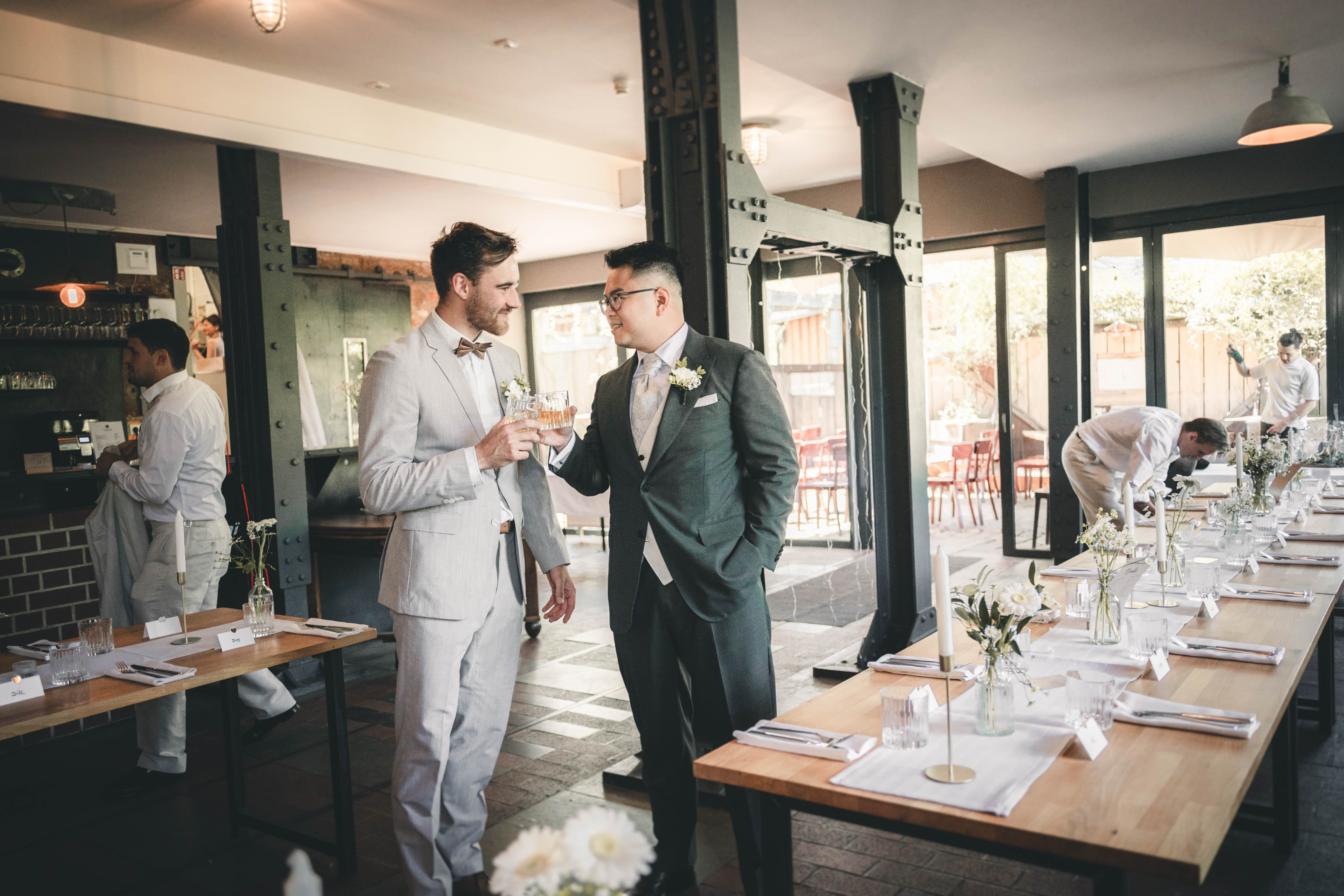 Die zwei Bräutigame stehen lächelnd mit ihren Drinks neben der elegant dekorierten Hochzeitstafel im modernen Industriesaal des Mörtelwerks Leipzig.