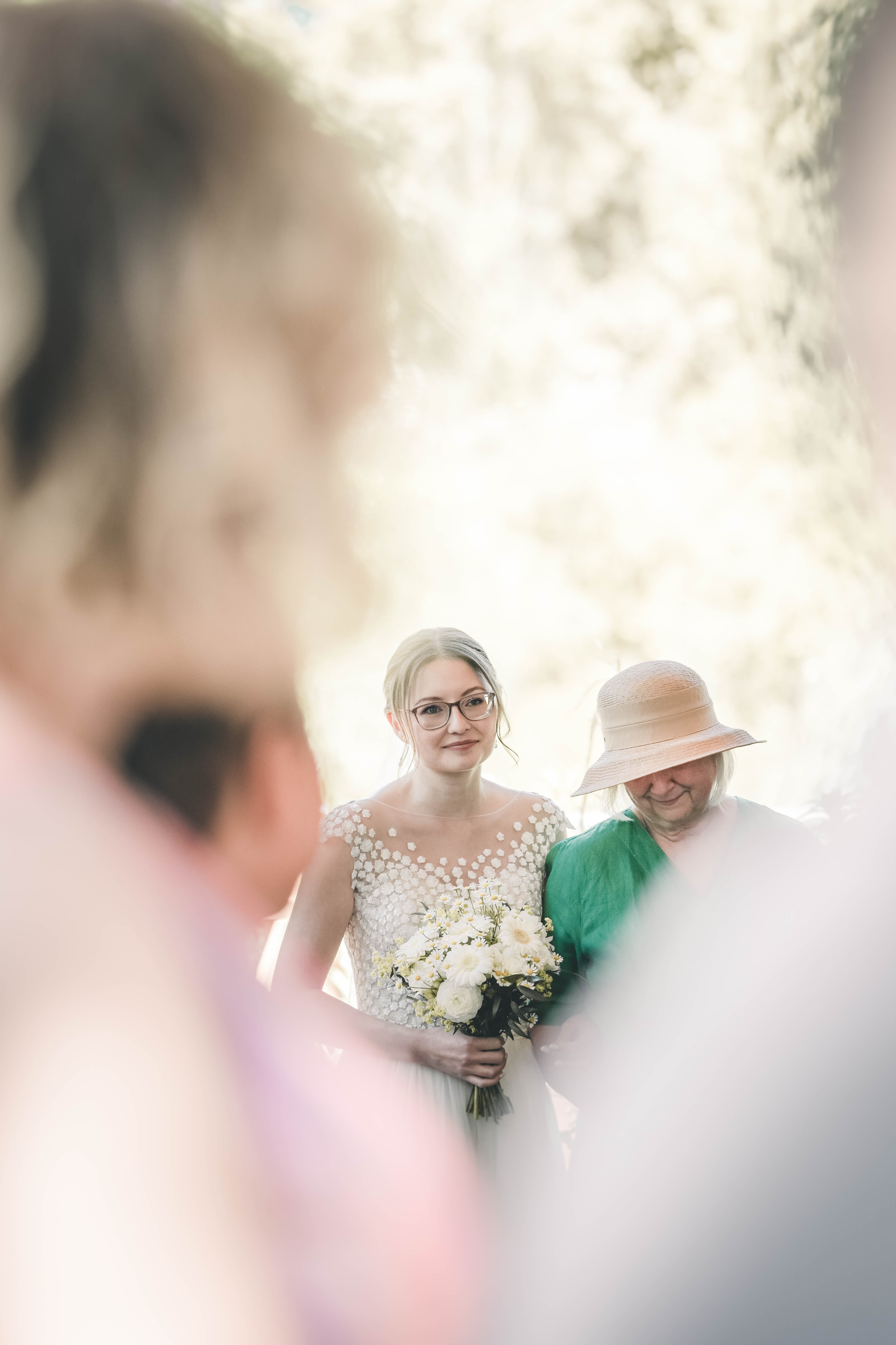 Ein fokussierter Blick auf die Braut im eleganten Kleid mit Wiesenblumenstrauß, die gerührt am Arm einer älteren Gästin mit Strohhut einzieht.