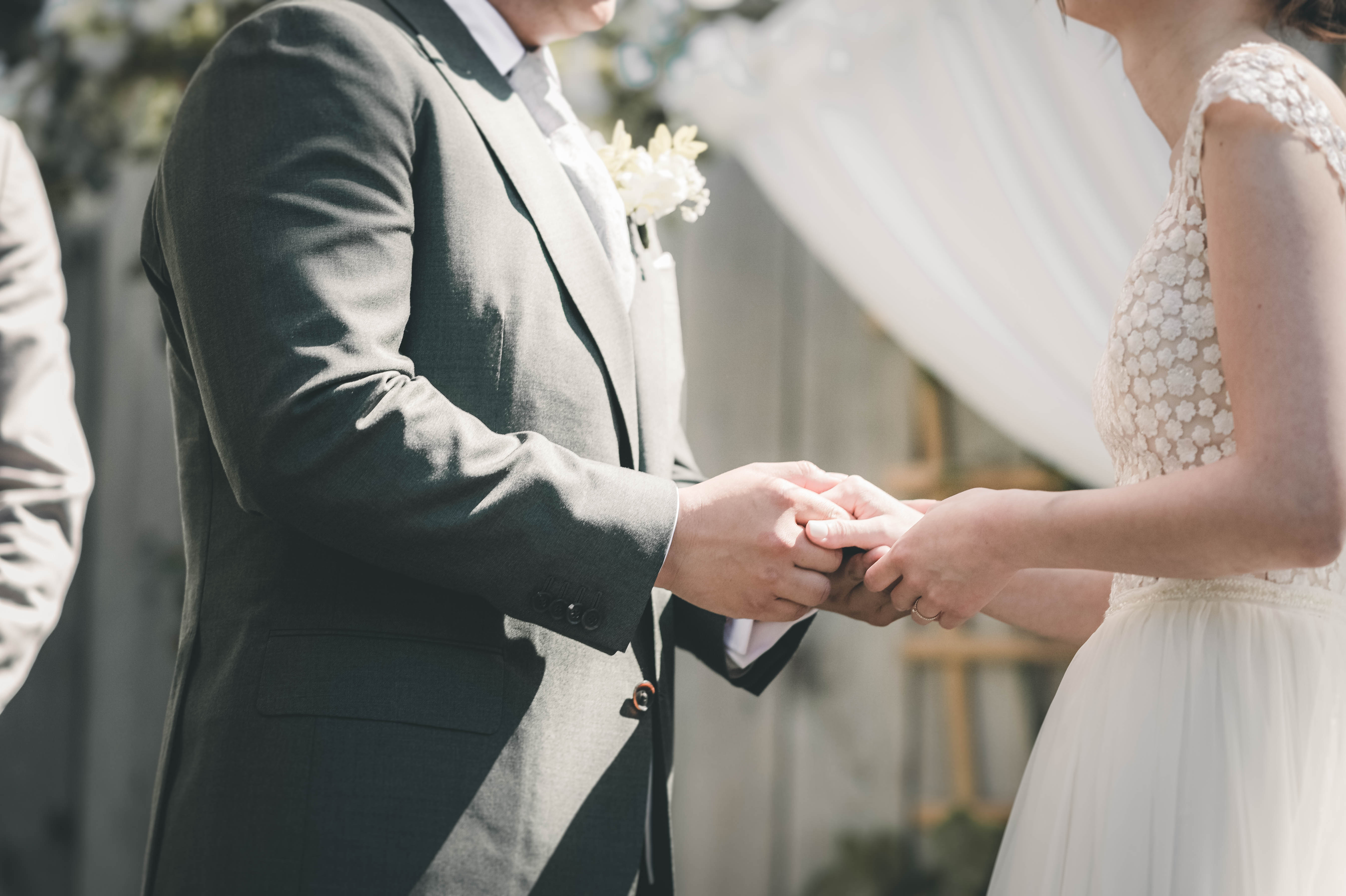 Eine detailreiche Nahaufnahme, wie sich die Braut im floralen Spitzenkleid und der Bräutigam im grauen Anzug während des Eheversprechens fest an den Händen halten.