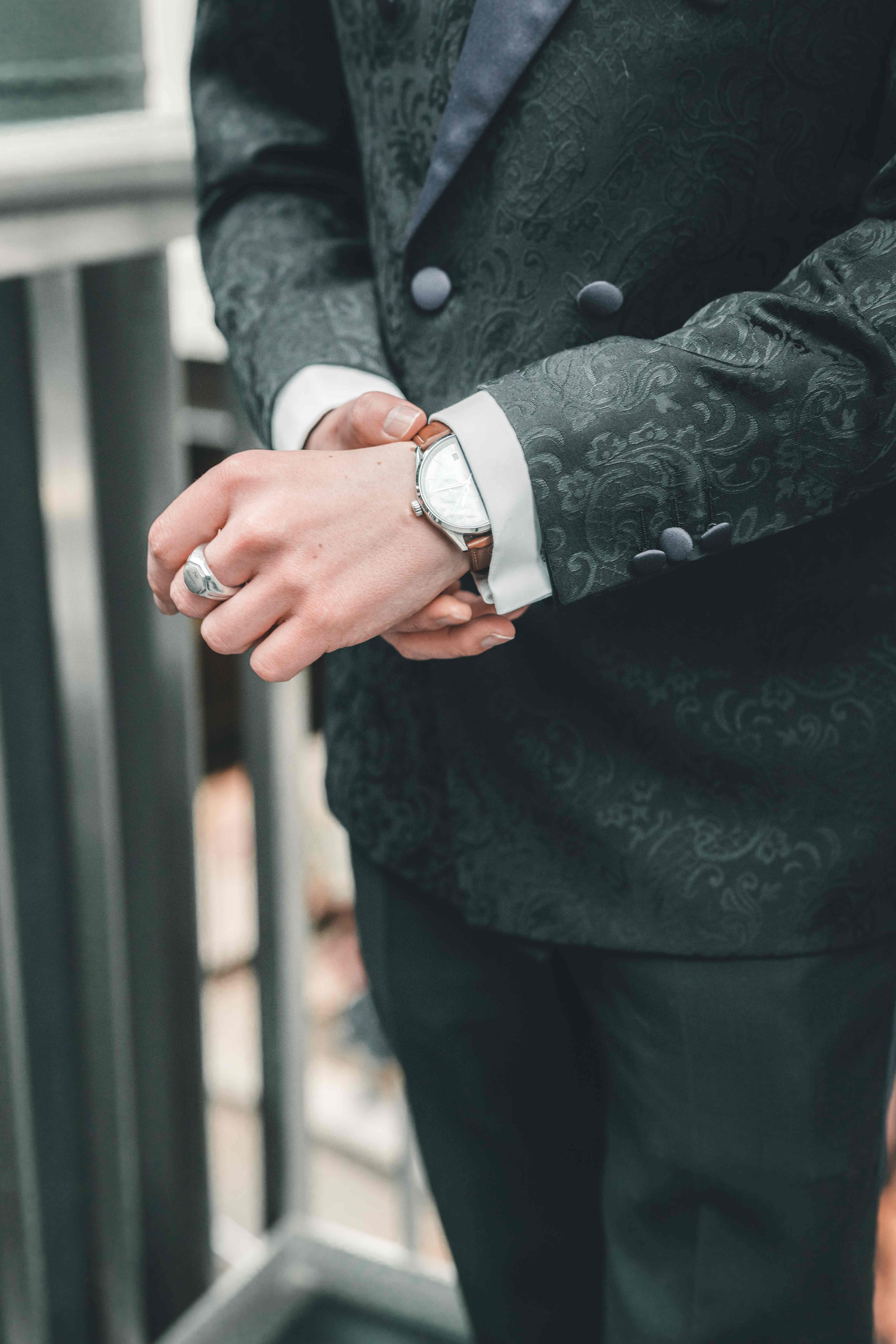 Nahaufnahme der Hände des Bräutigams im edel gemusterten Hochzeitsanzug, der seine Armbanduhr mit braunem Lederband und einen Siegelring zeigt.