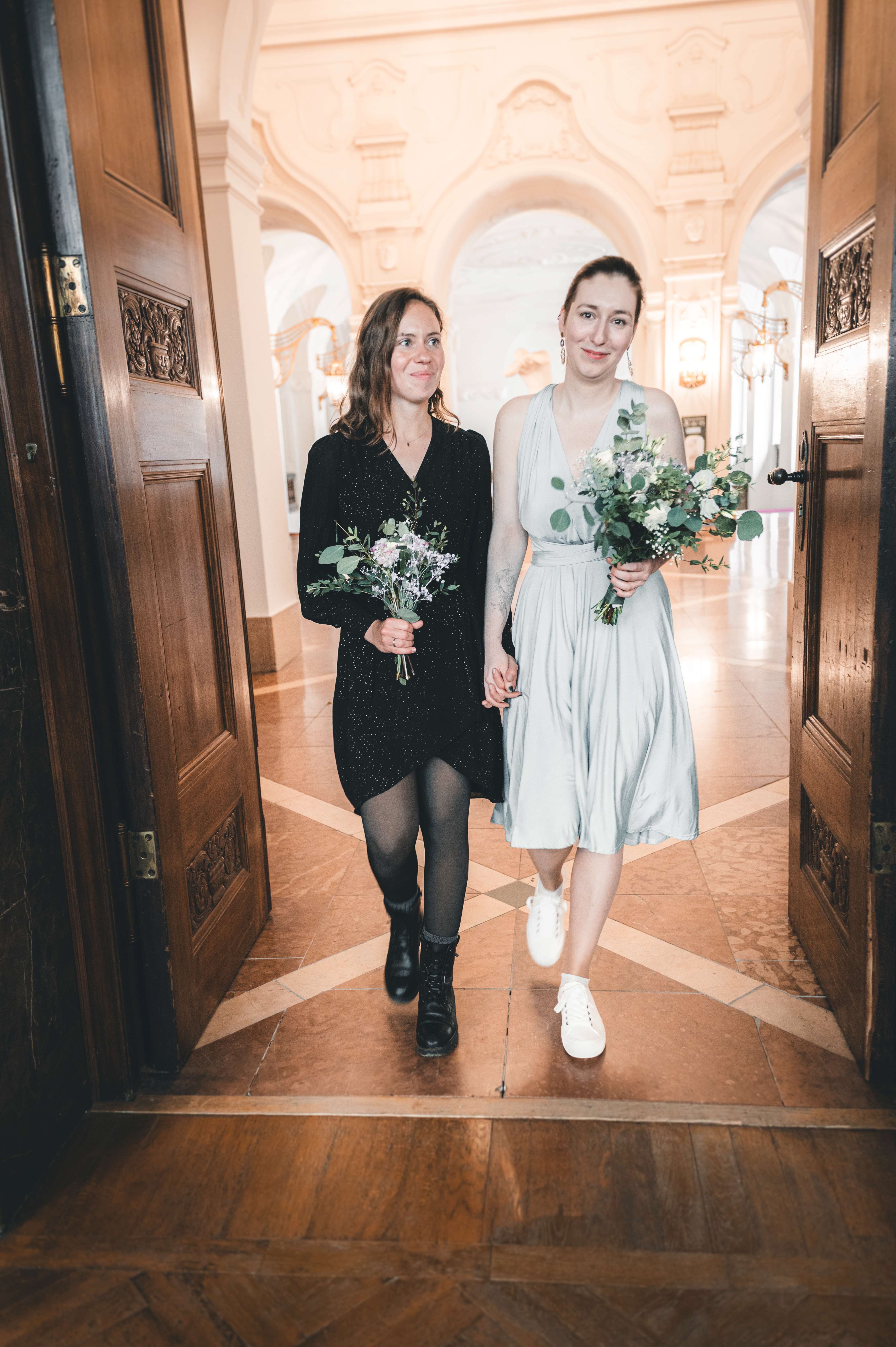 Braut und Brautjungfer im eleganten schwarzen und salbeigrünen Kleid schreiten Hand in Hand durch die geöffneten Holzflügeltüren in den Hochzeitssaal.