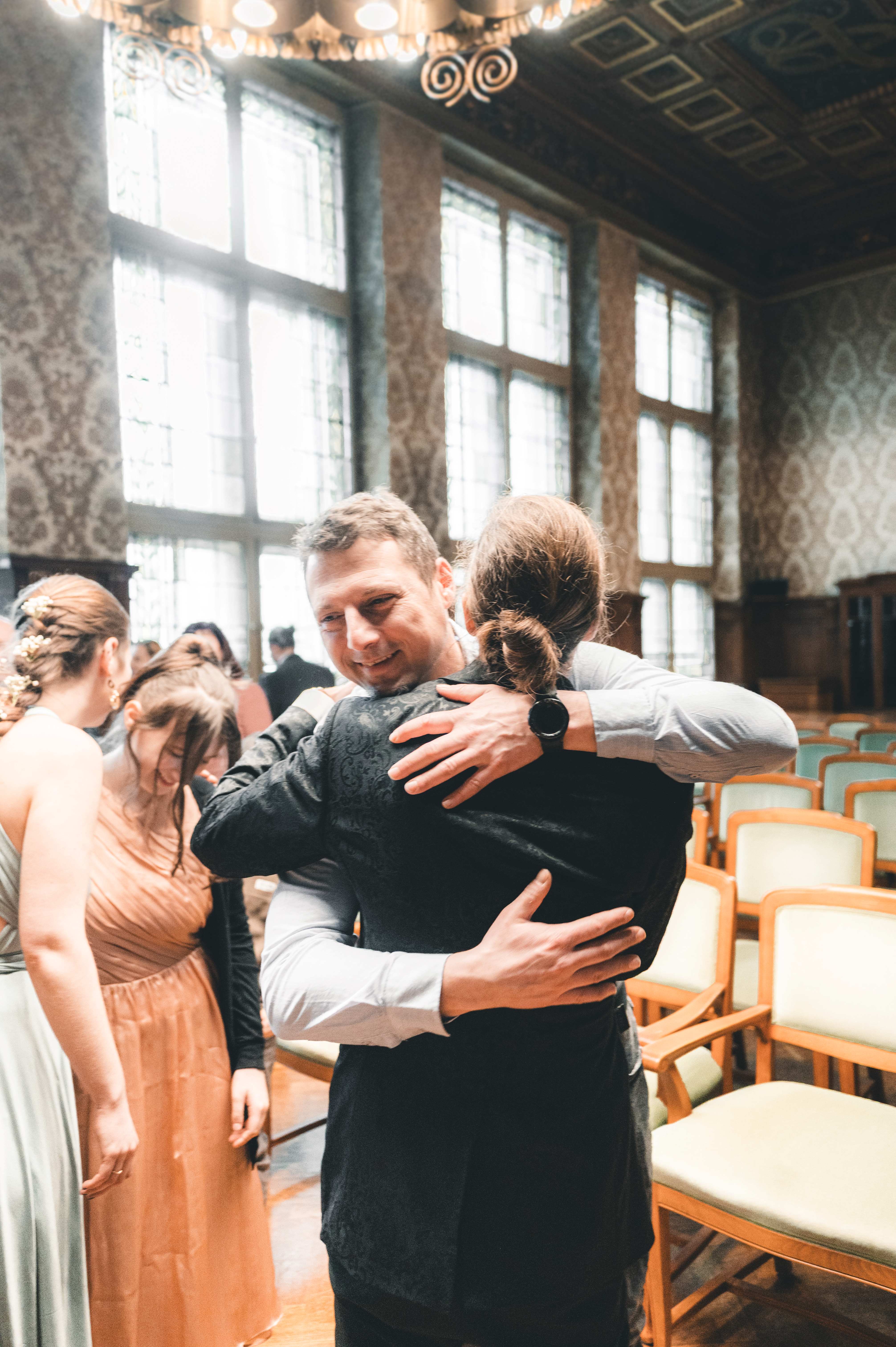 Ein männlicher Hochzeitsgast umarmt den Bräutigam im Ratsplenarsaal Leipzig herzlich zur Gratulation, während die Braut daneben steht.