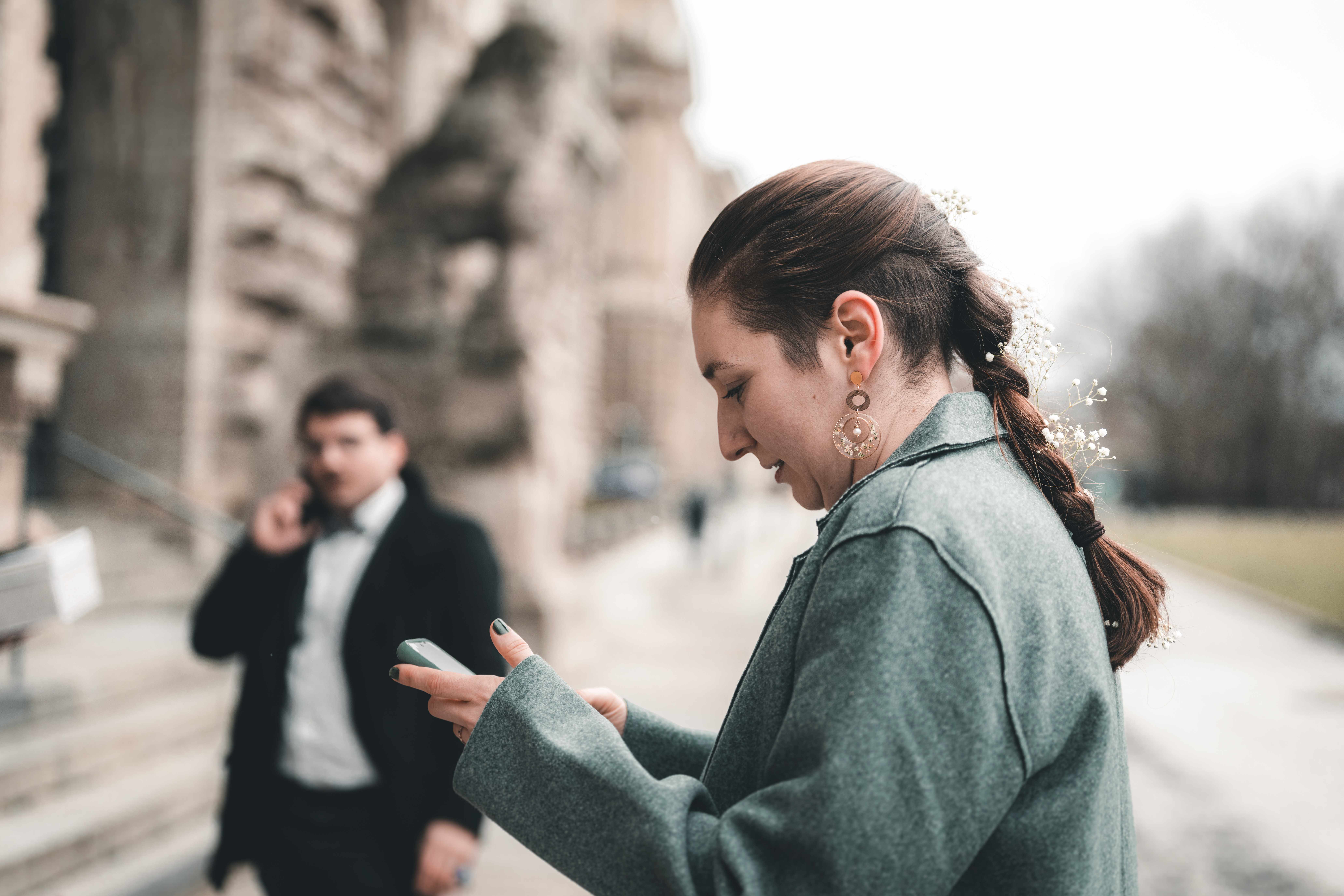 Die Braut im grünen Mantel blickt vor dem Neuen Rathaus auf ihr Smartphone, während im Hintergrund ein Hochzeitsgast im Anzug unscharf zu sehen ist.
