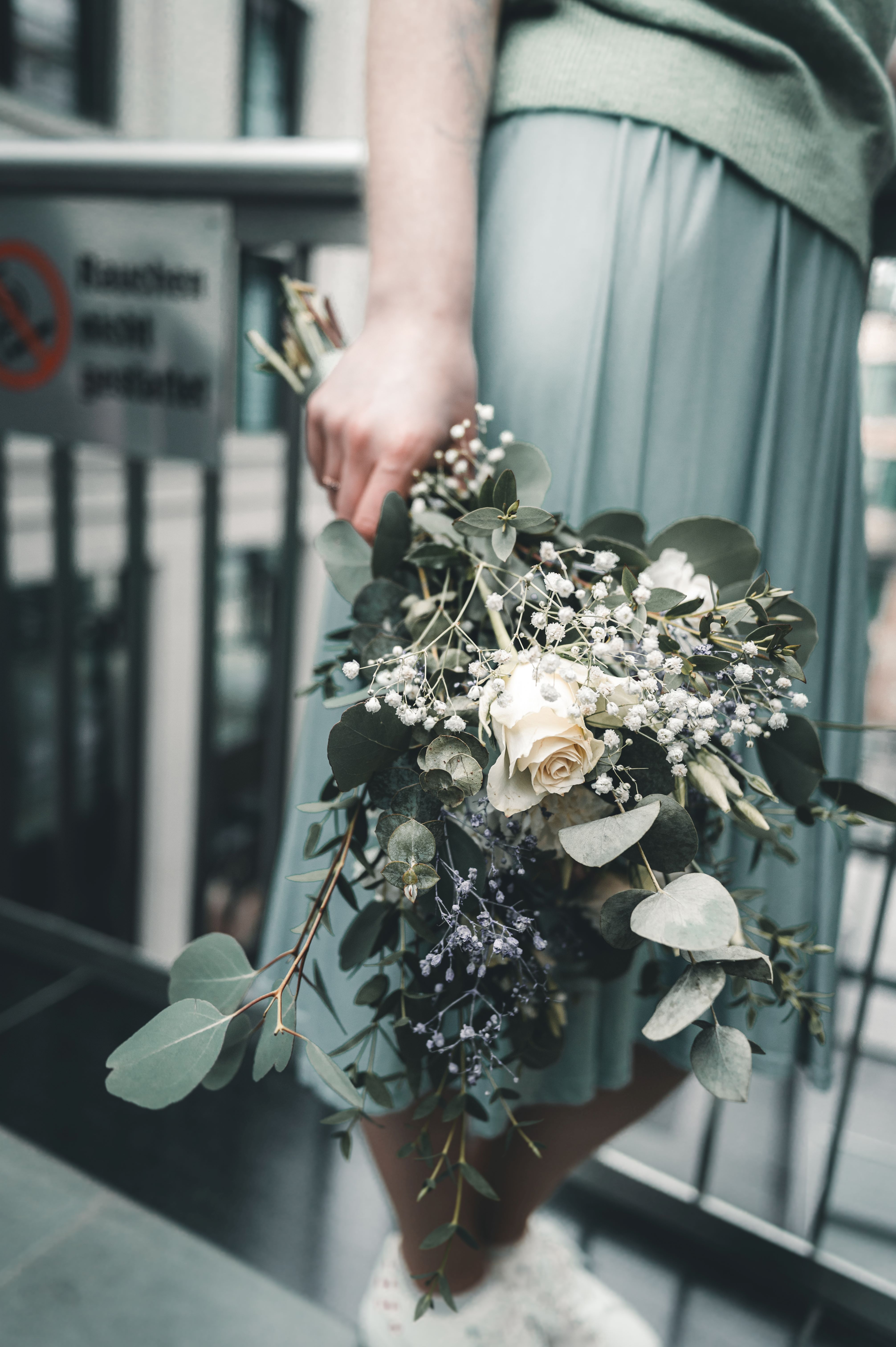 Nahaufnahme des lockeren Brautstraußes aus weißen Rosen, Schleierkraut und Eukalyptus, den die Braut lässig an ihrer Seite hält.
