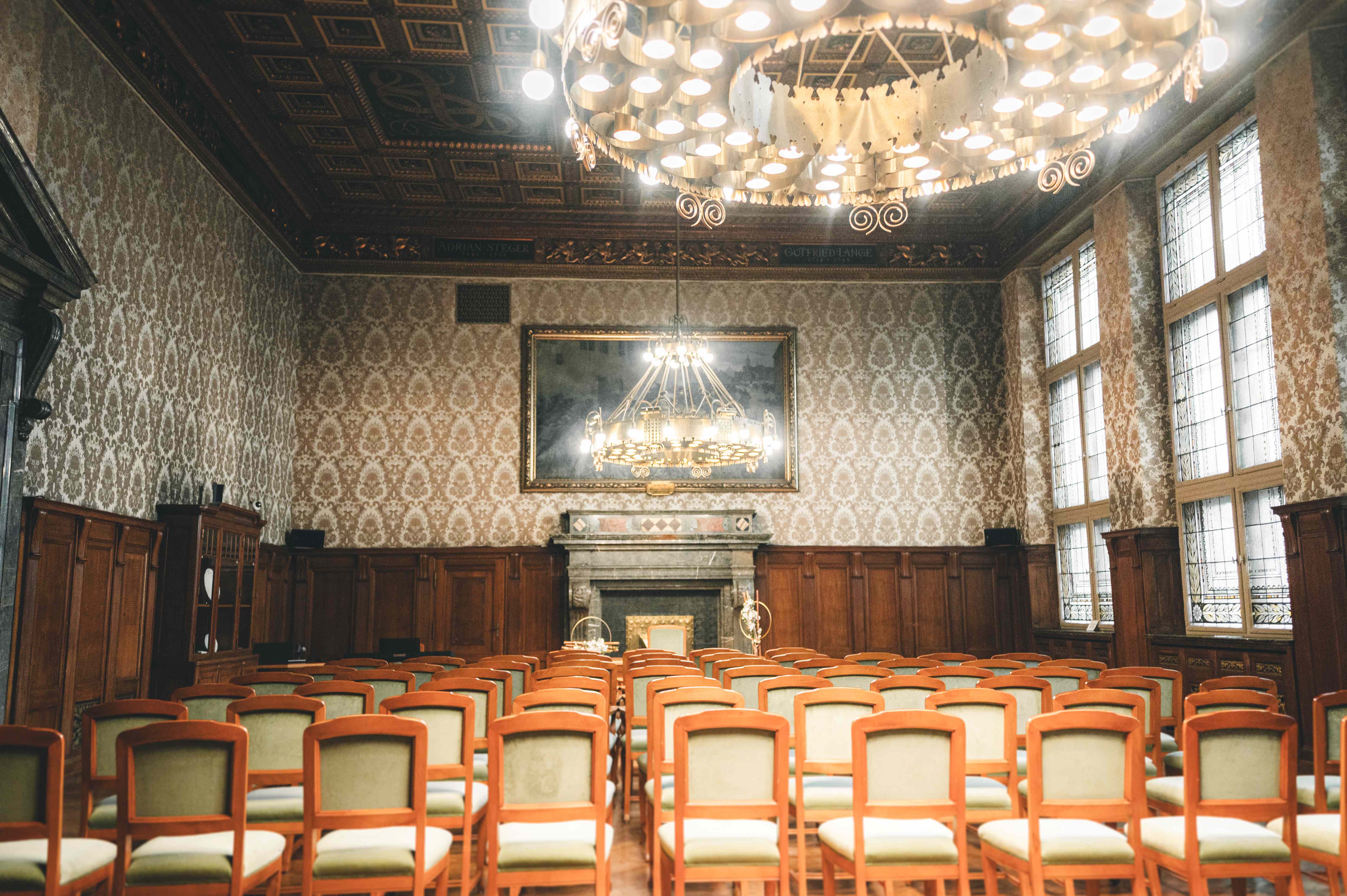 Die Innenansicht des Ratsplenarsaals in Leipzig mit langen Reihen von Holzstühlen, prunkvollen Tapeten und einem großen historischen Gemälde an der Stirnseite.