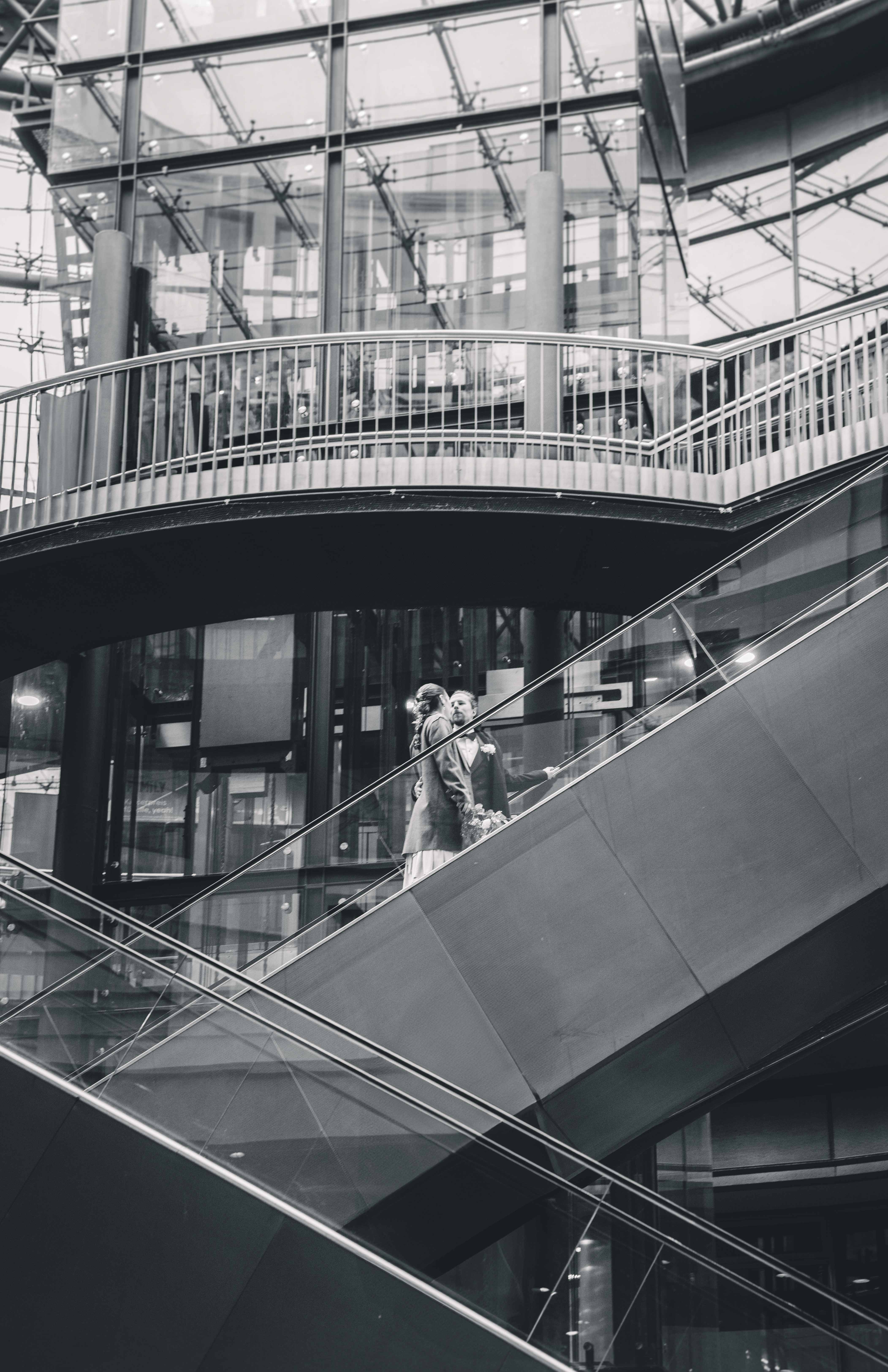 Eine architektonisch ansprechende Schwarz-Weiß-Aufnahme des Paares, das auf einer Rolltreppe inmitten der modernen Glas- und Stahlkonstruktion der Strohsack-Passage steht.