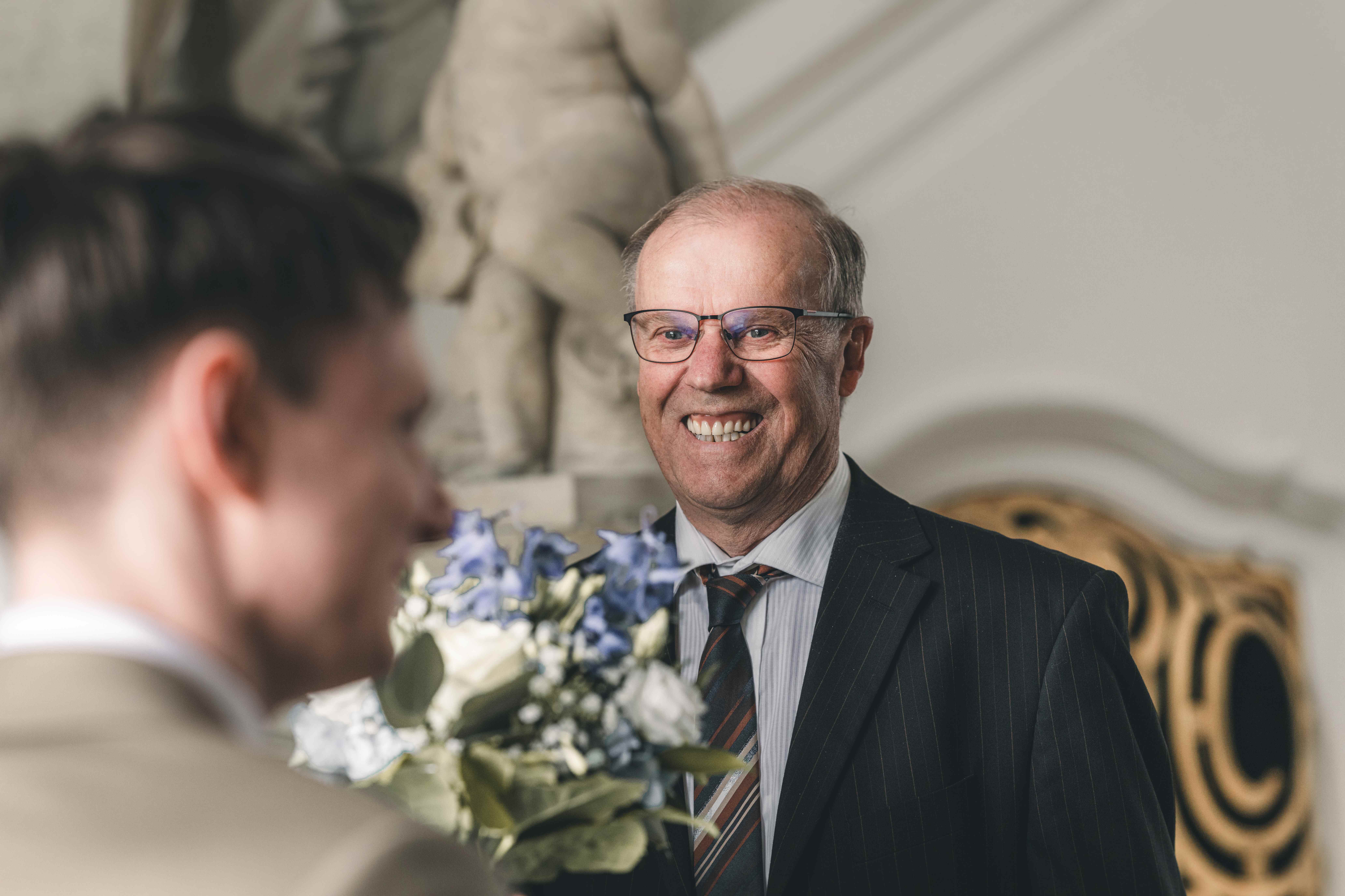 Ein älterer Hochzeitsgast oder der Bräutigamvater im eleganten Nadelstreifenanzug blickt den Bräutigam im Trausaal mit einem stolzen, herzlichen Lächeln an.