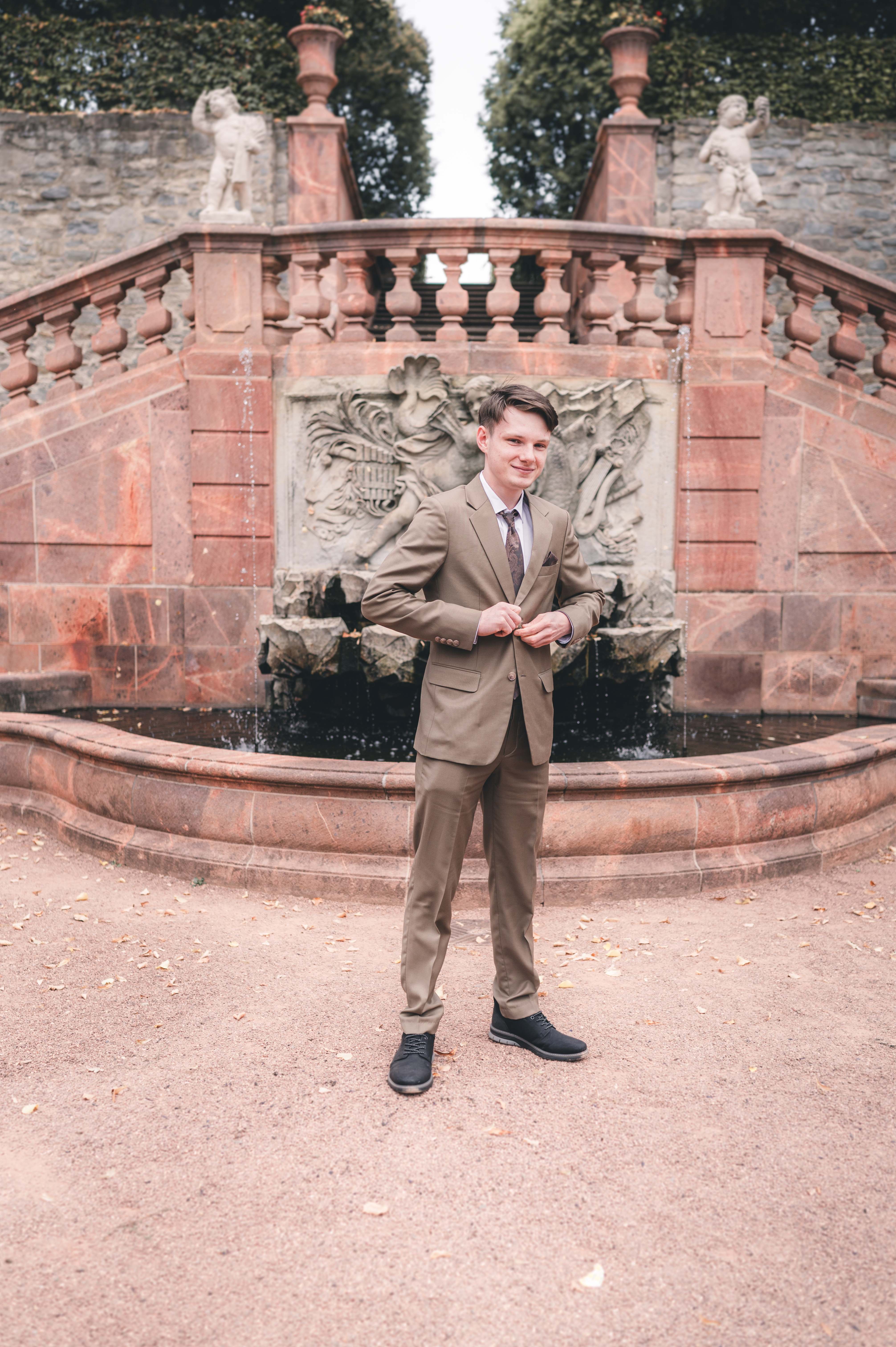 Der Bräutigam posiert im modernen hellbraunen Hochzeitsanzug lächelnd vor den Wasserspielen im Schlosspark Lichtenwalde nahe Chemnitz.