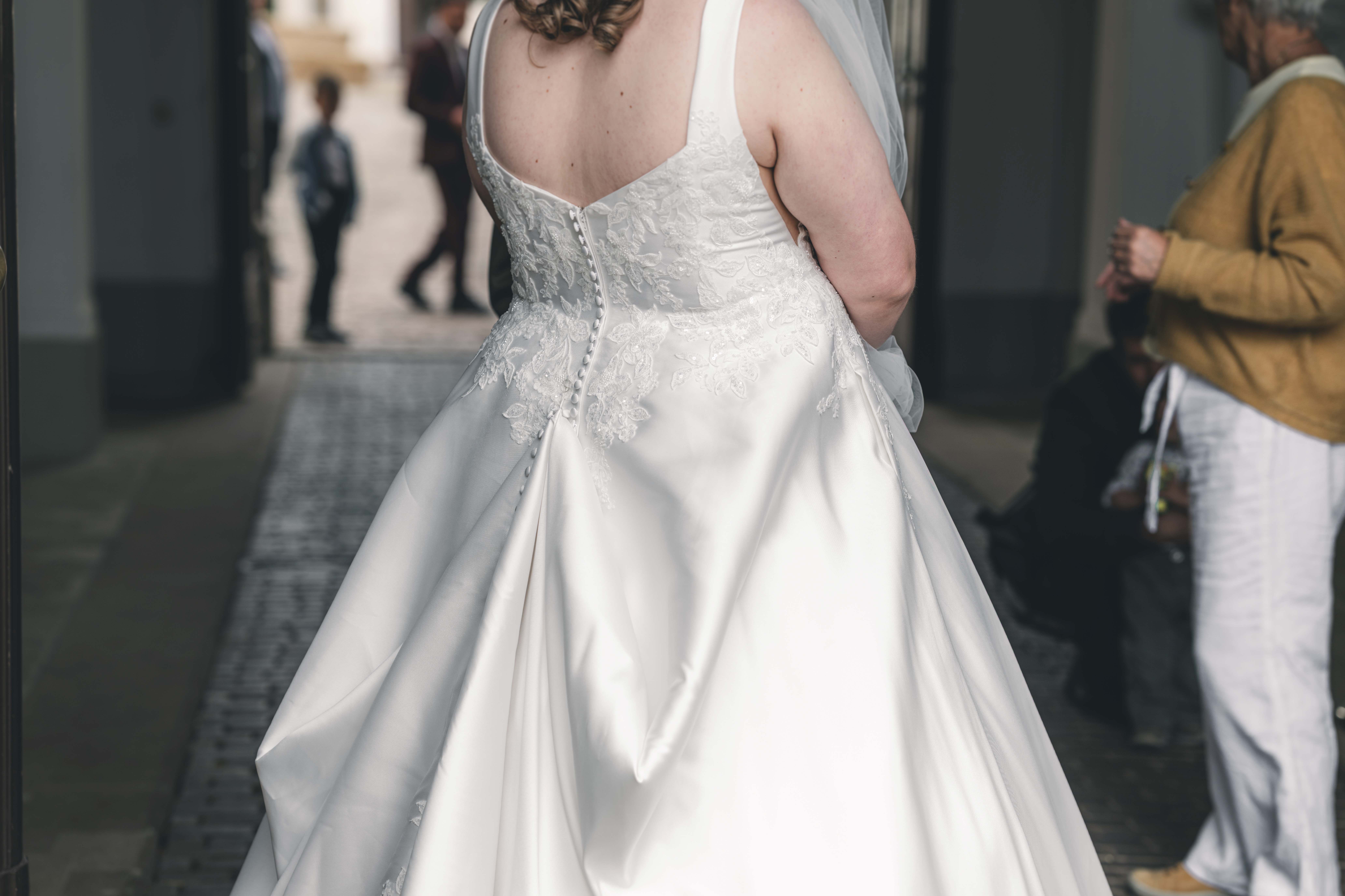 Rückansicht des eleganten Brautkleides beim Einzug in den Saal, mit einer feinen Knopfleiste und edler floraler Spitze am Rücken.