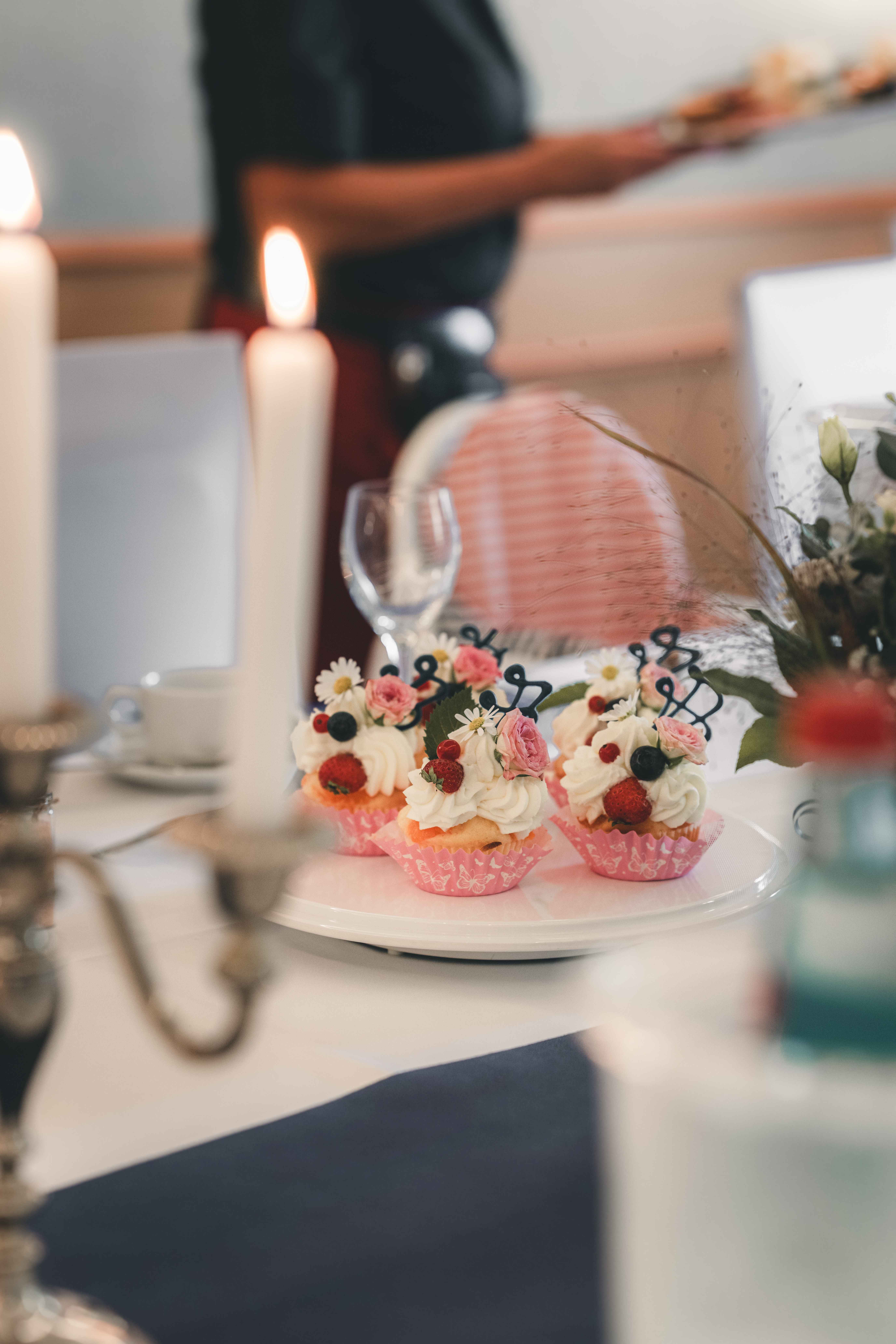 Detailaufnahme von süßen Hochzeits-Cupcakes mit weißer Cremehaube, frischen Beeren und kleinen echten Rosen auf dem festlichen Tisch im Kerzenschein.