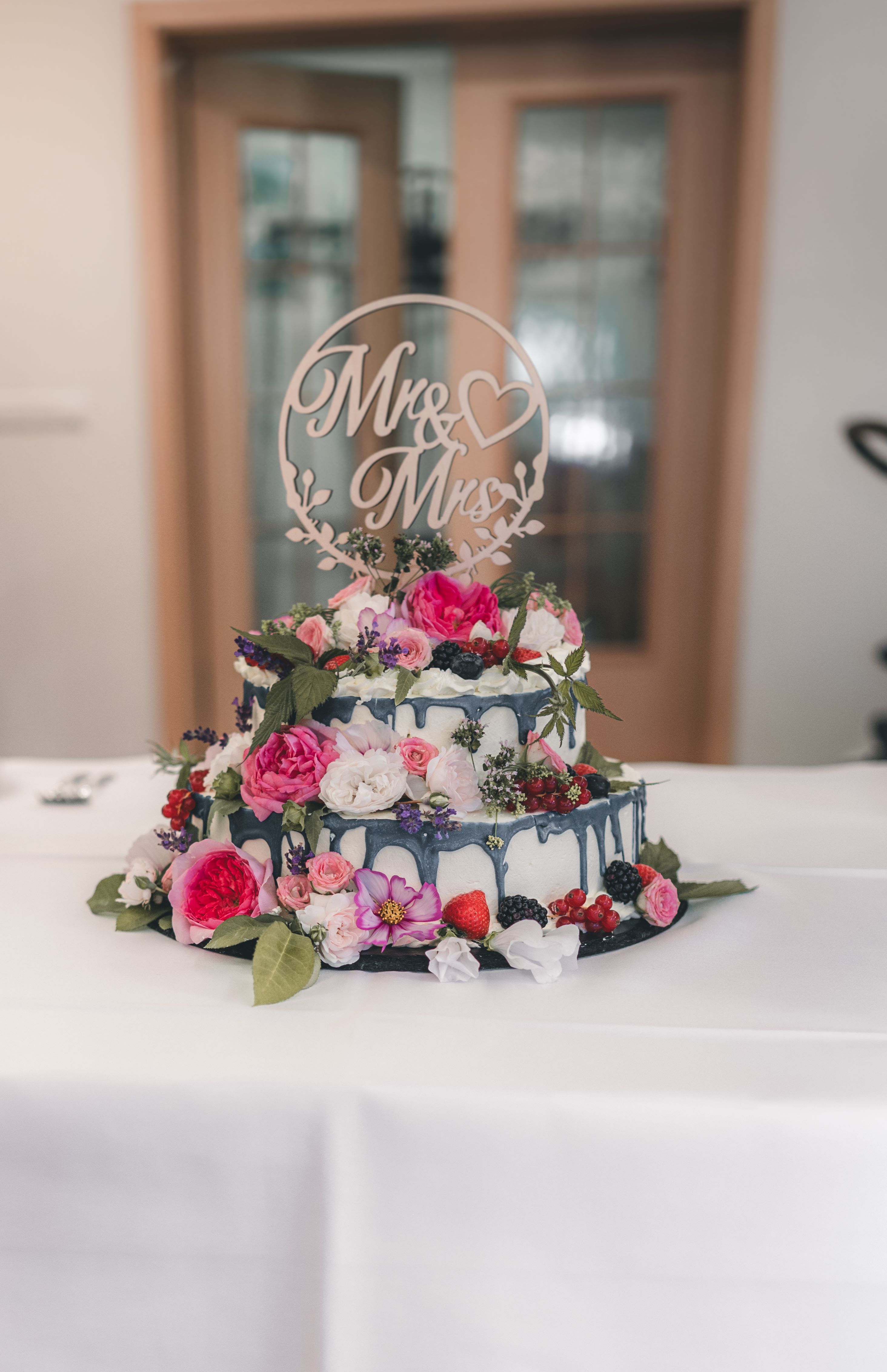 Eine zweistöckige weiße Torte mit blauem Drip-Effekt, üppig dekoriert mit frischen Beeren, Rosen und einem hölzernen „Mr & Mrs“-Schriftzug.