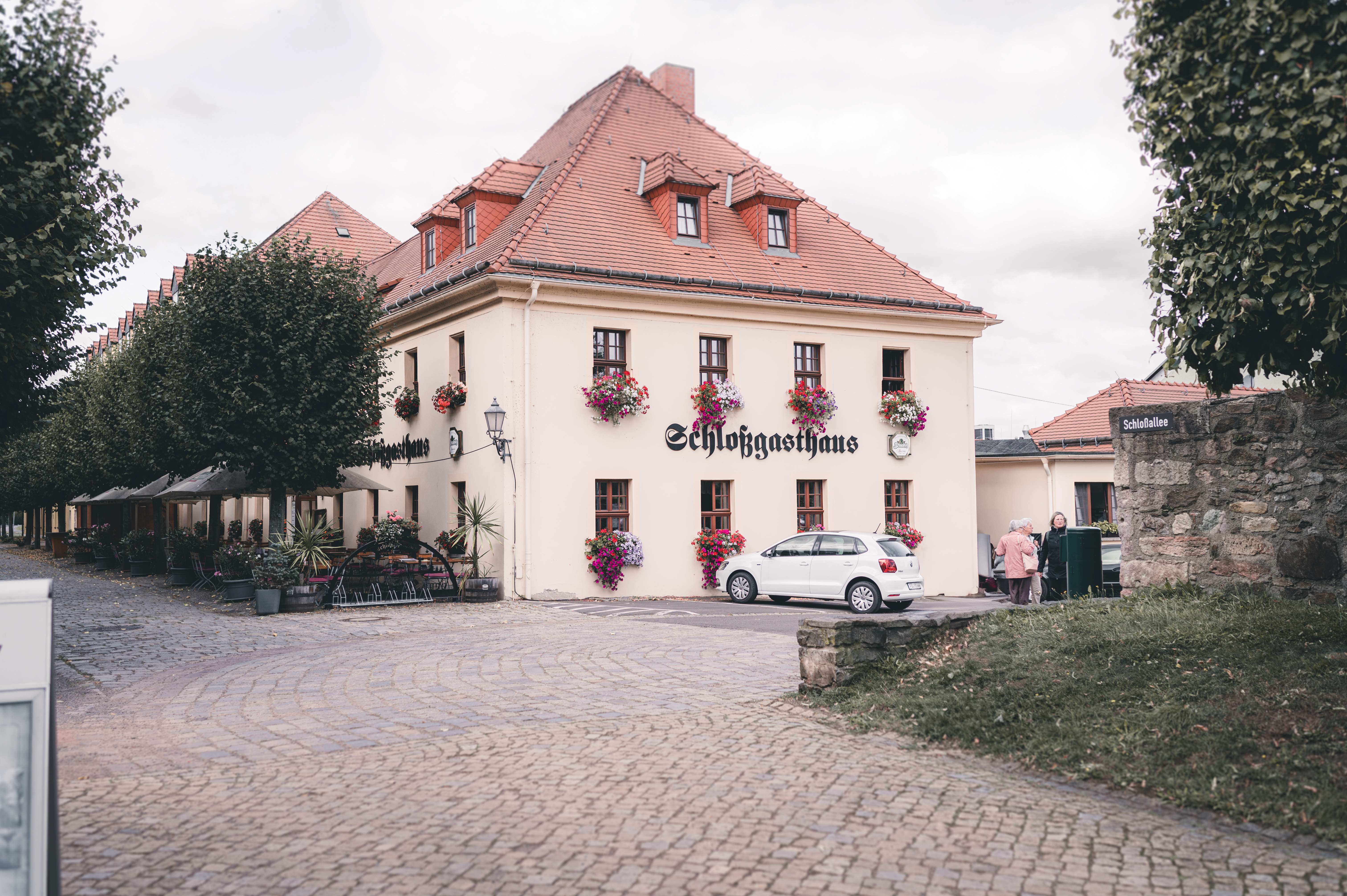 Die Außenansicht des hellen Schlossgasthauses in Lichtenwalde bei Chemnitz mit weiten Pflasterwegen und einladenden Sonnenschirmen im Sommer.