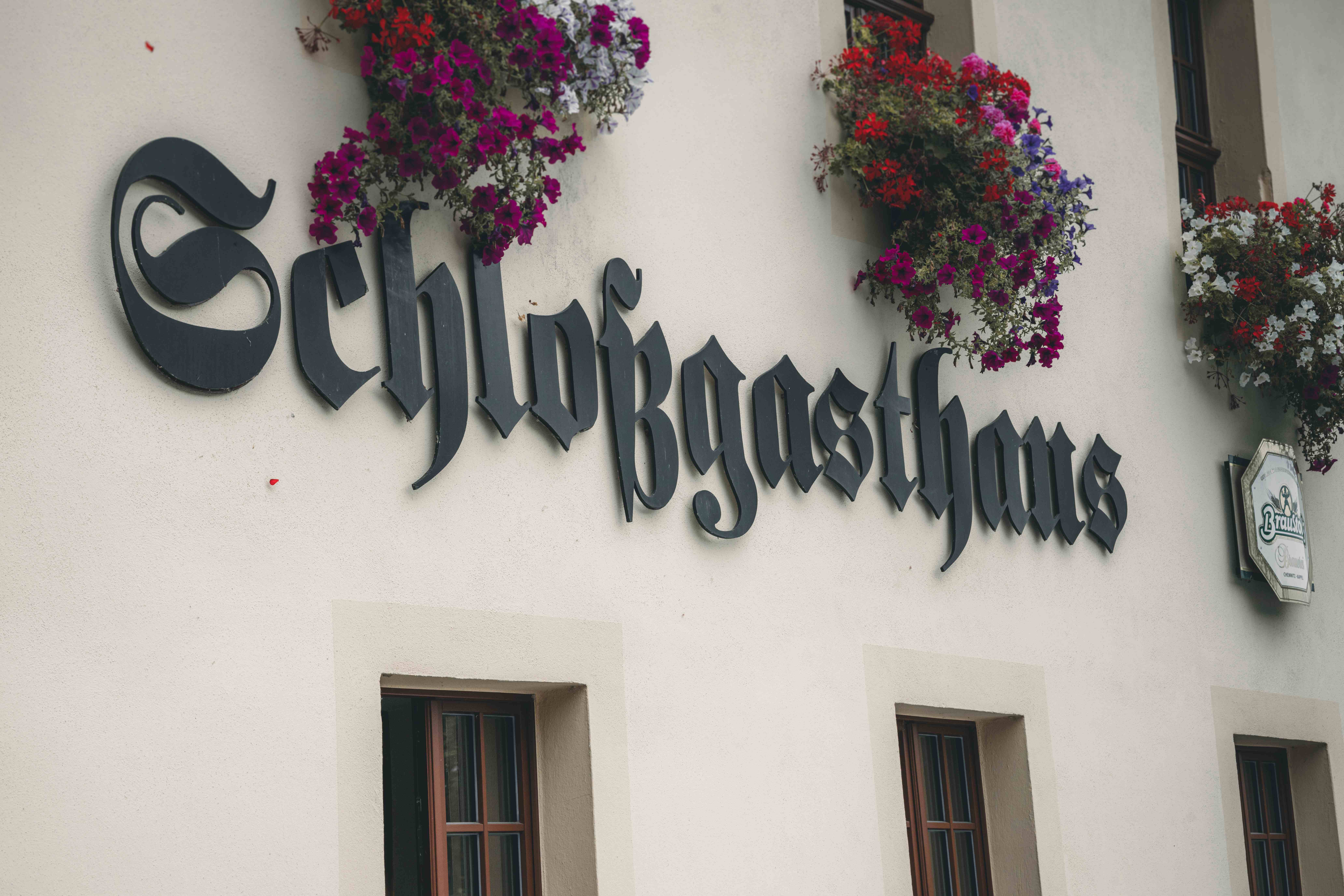 Nahaufnahme des geschwungenen, schwarzen Schriftzugs „Schlossgasthaus“ an eurer Hochzeitslocation, verziert mit bunten Blumenkästen.