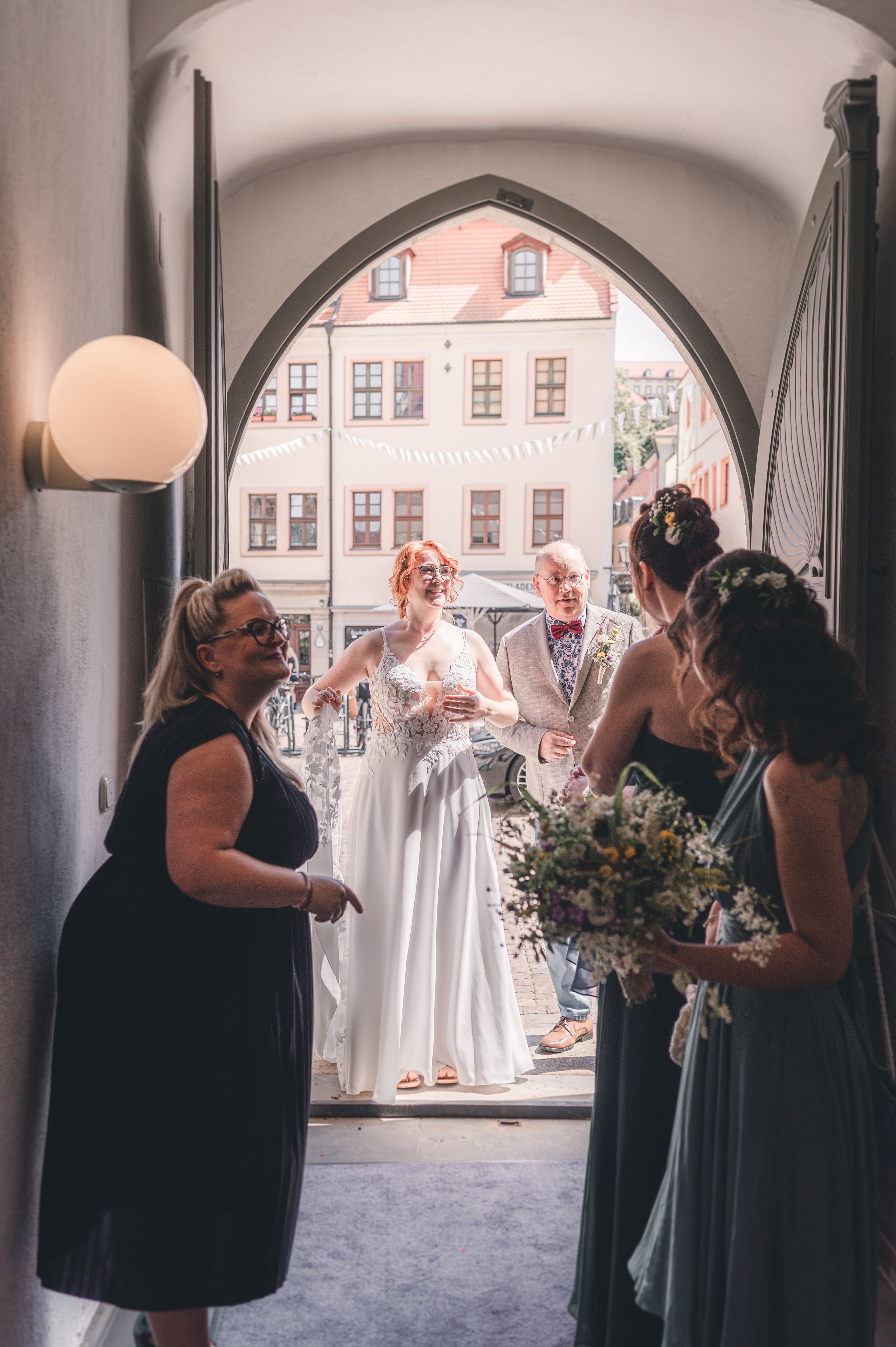 Die rothaarige Braut im eleganten Spitzenkleid betritt in Begleitung ihres Vaters das historische Portal des Standesamtes Pirna, während die Brautjungfern warten.