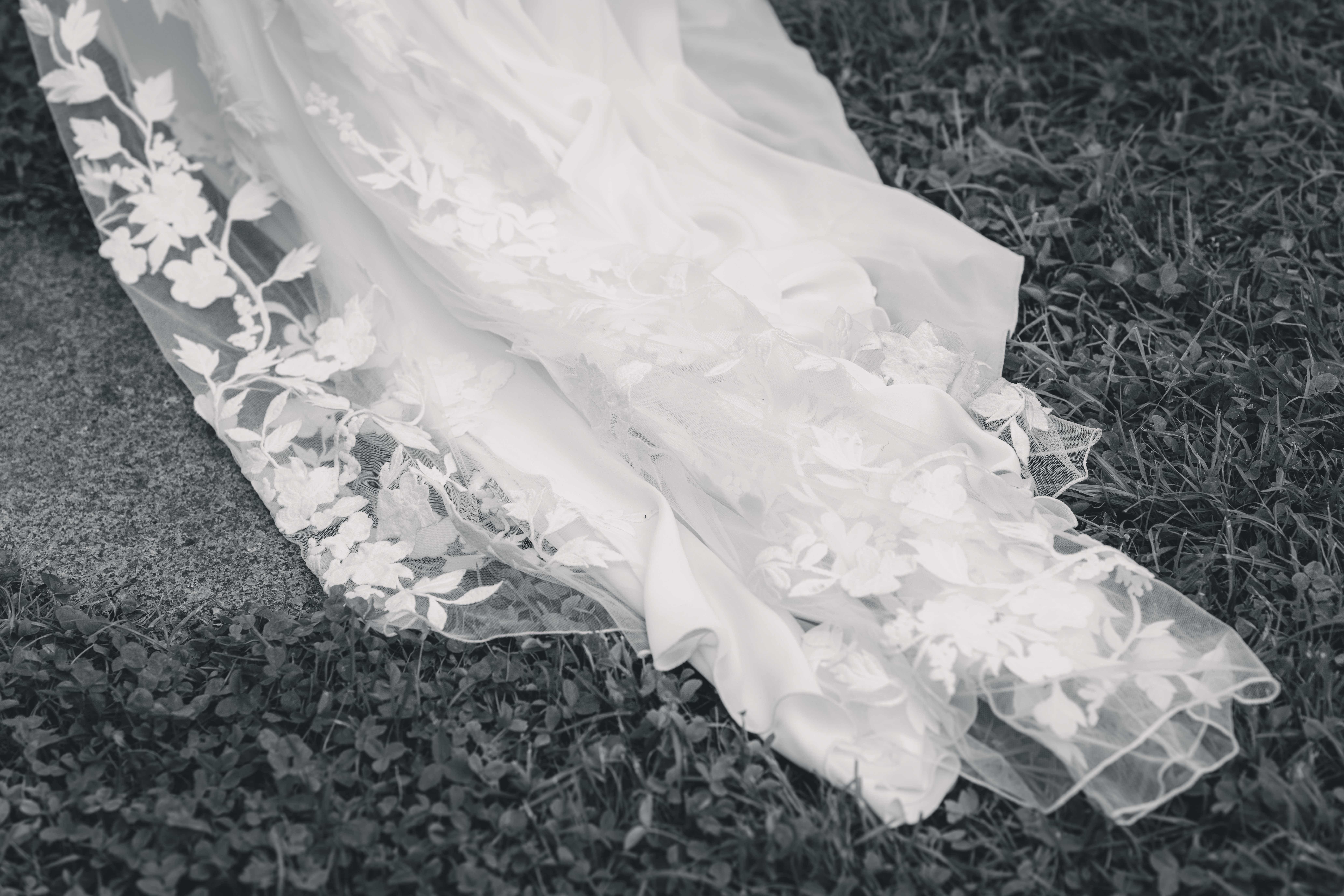 Eine Schwarz-Weiß-Nahaufnahme der eleganten Brautkleidschleppe mit floralen Spitzen-Applikationen auf dem Rasen in der Oberlausitz.