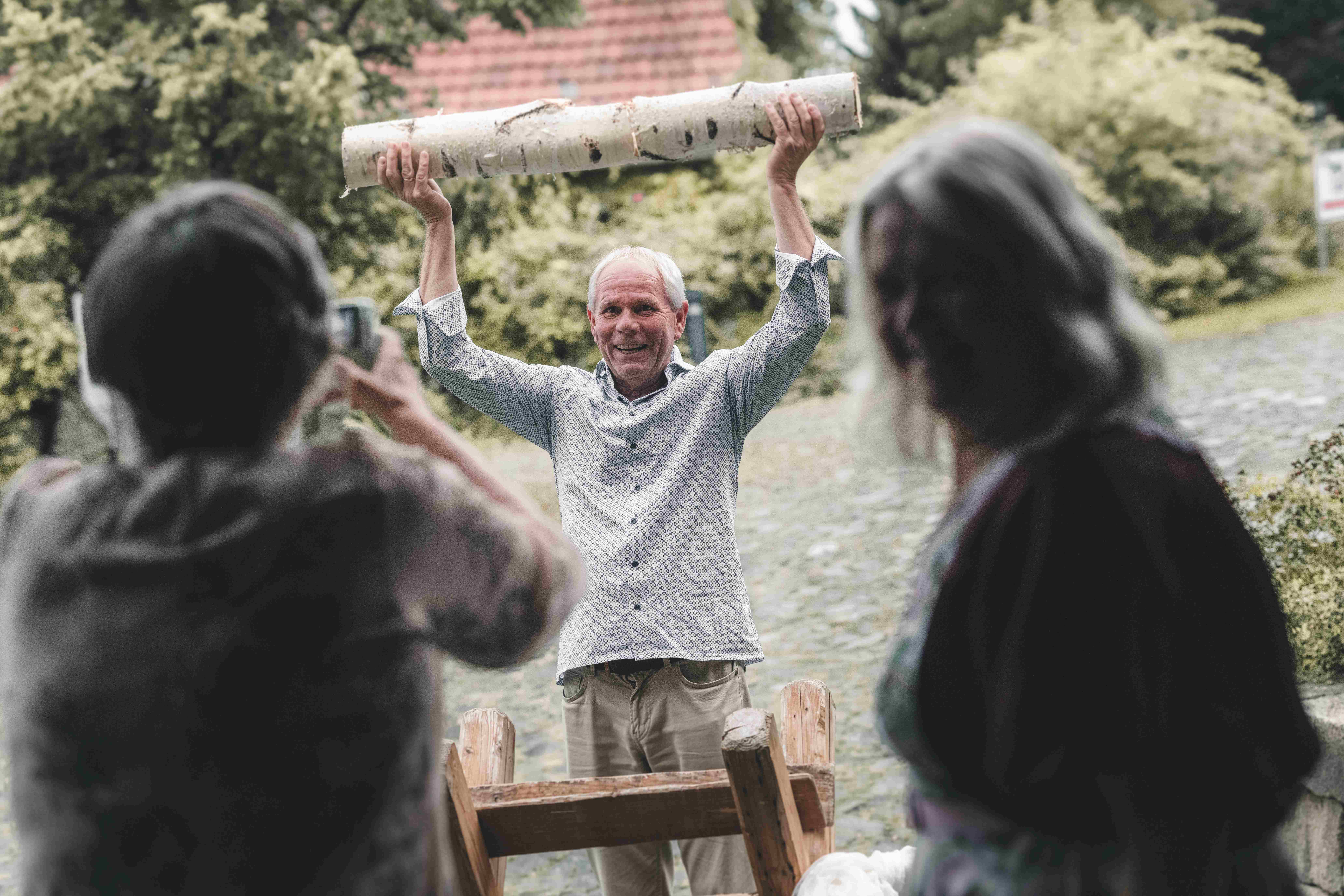 Ein lachender Hochzeitsgast hebt den frisch durchgesägten Birkenstamm triumphierend in die Höhe, während das Brautpaar applaudiert.
