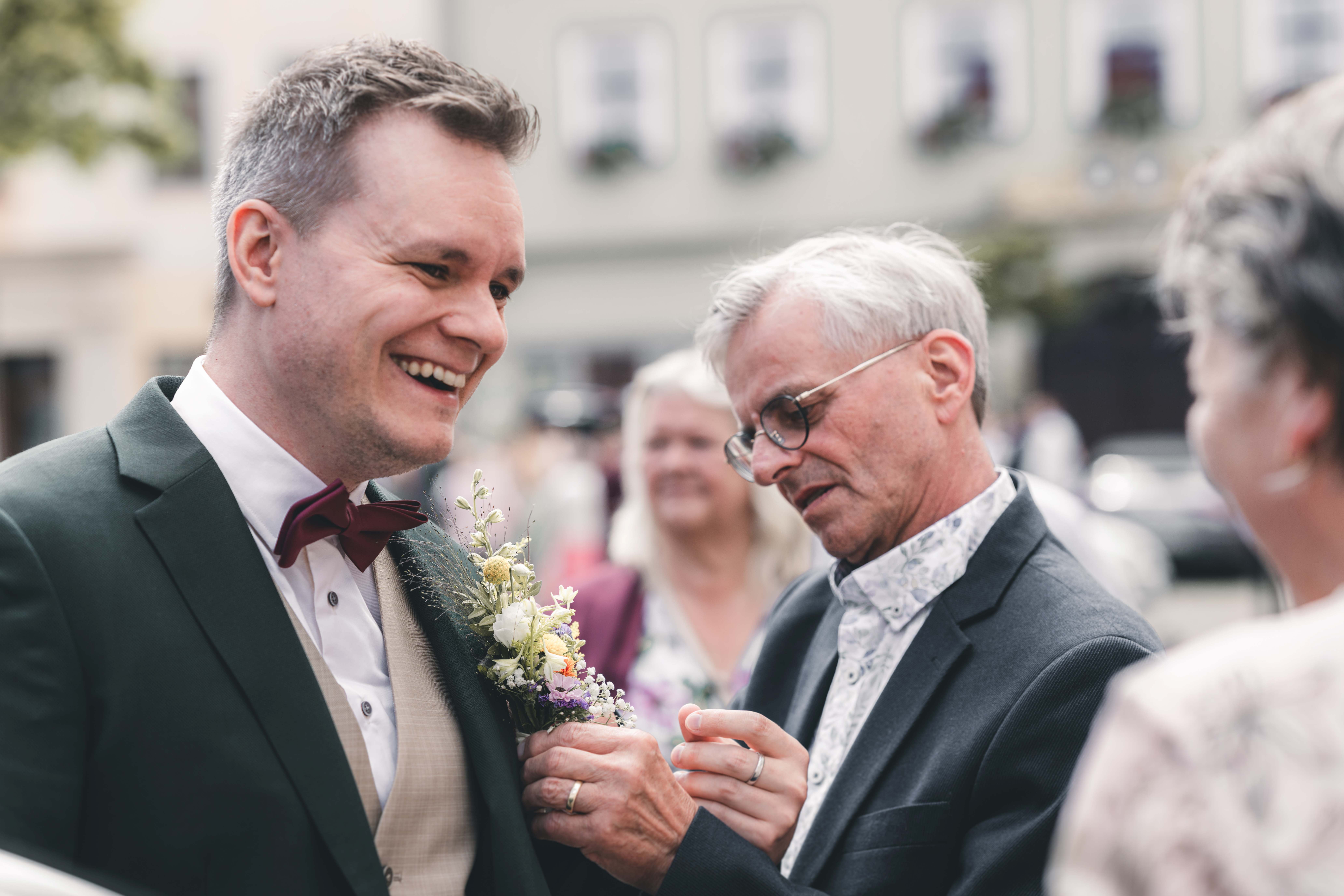 Ein älterer Gast befestigt dem glücklich lächelnden Bräutigam den floralen Ansteckstrauß aus Wiesenblumen am Anzug vor der Trauung.
