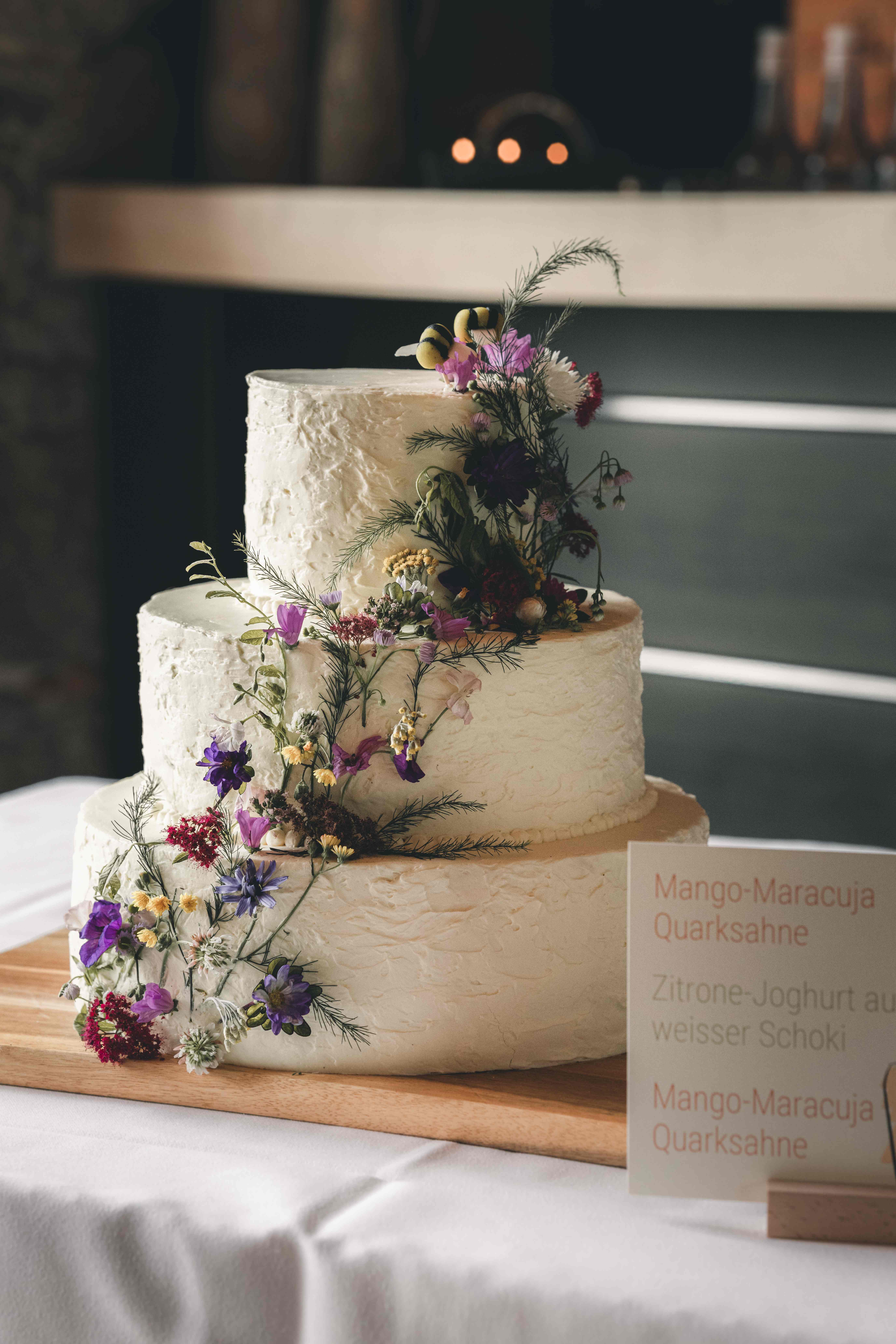 Eine dreistöckige, weiße Hochzeitstorte im "Semi-Naked"-Stil, kunstvoll dekoriert mit bunten Wiesenblumen auf einem Holzbrett in Oppach.