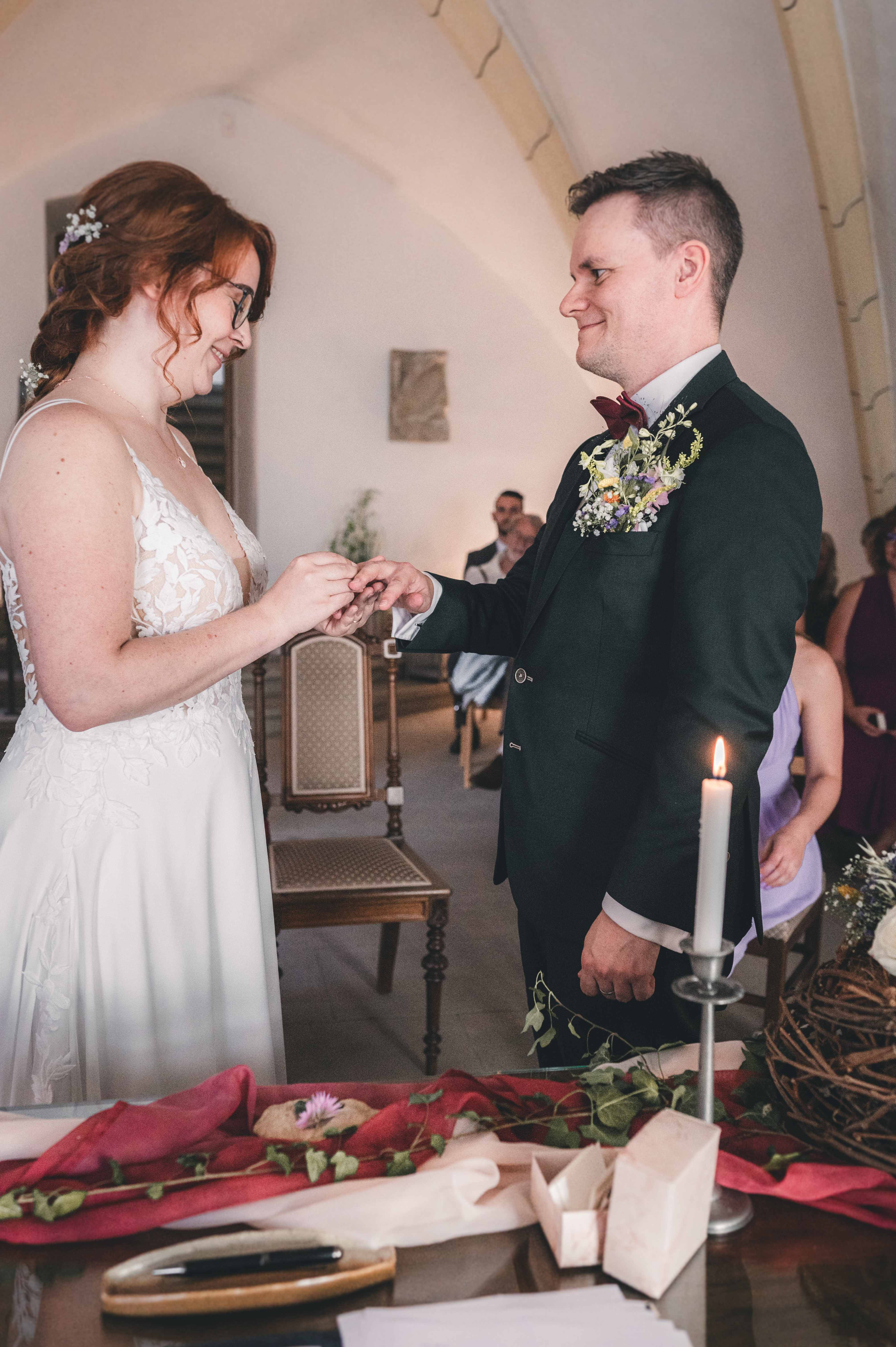 Die Braut steckt ihrem Bräutigam lächelnd den Ehering an den Finger, im Vordergrund brennt eine Hochzeitskerze.