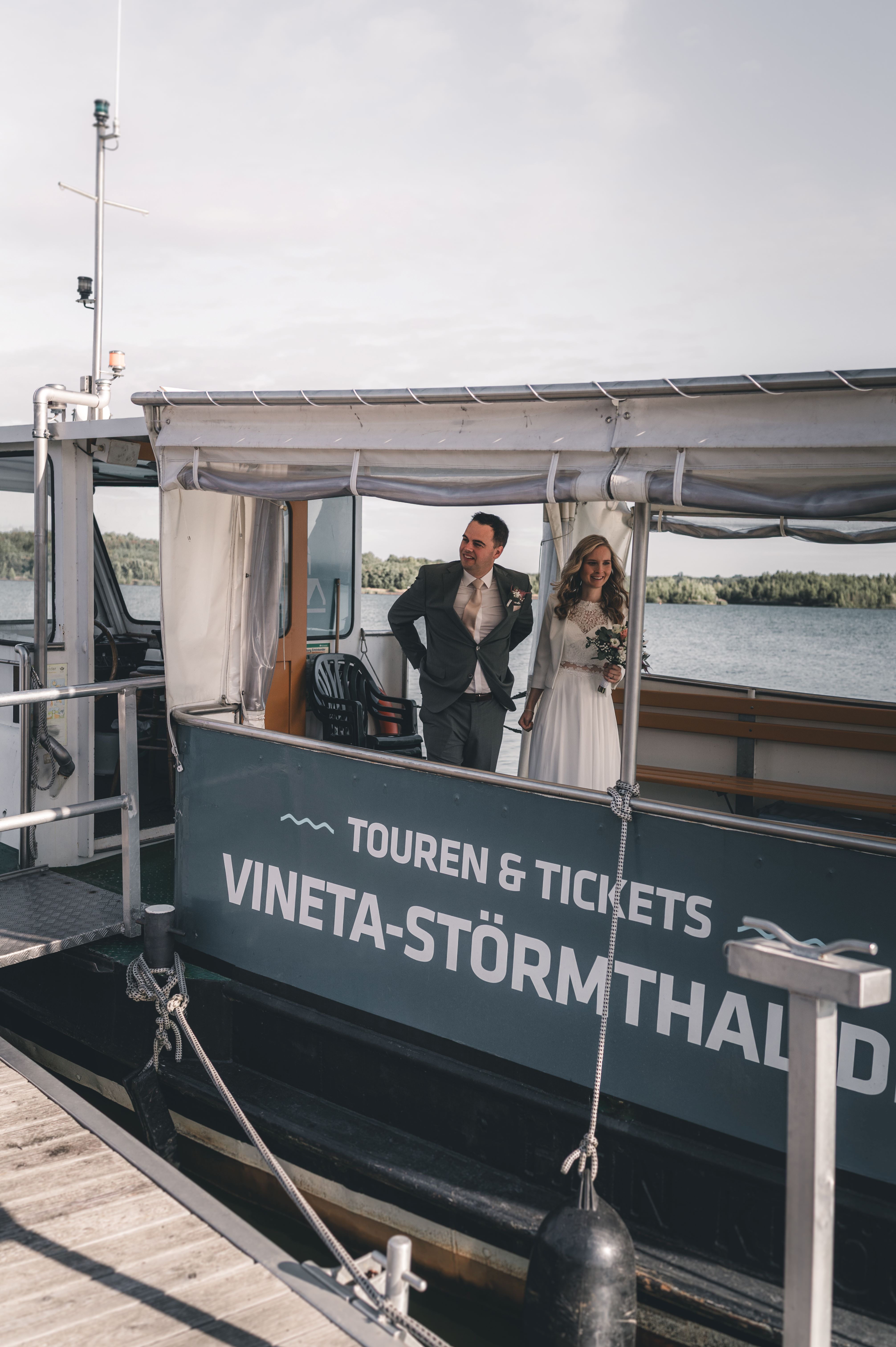 Das Brautpaar steht gut gelaunt an Deck des Schiffes mit der Aufschrift „VINETA-STÖRMTHALER SEE“ während der Fahrt zur Trauung.