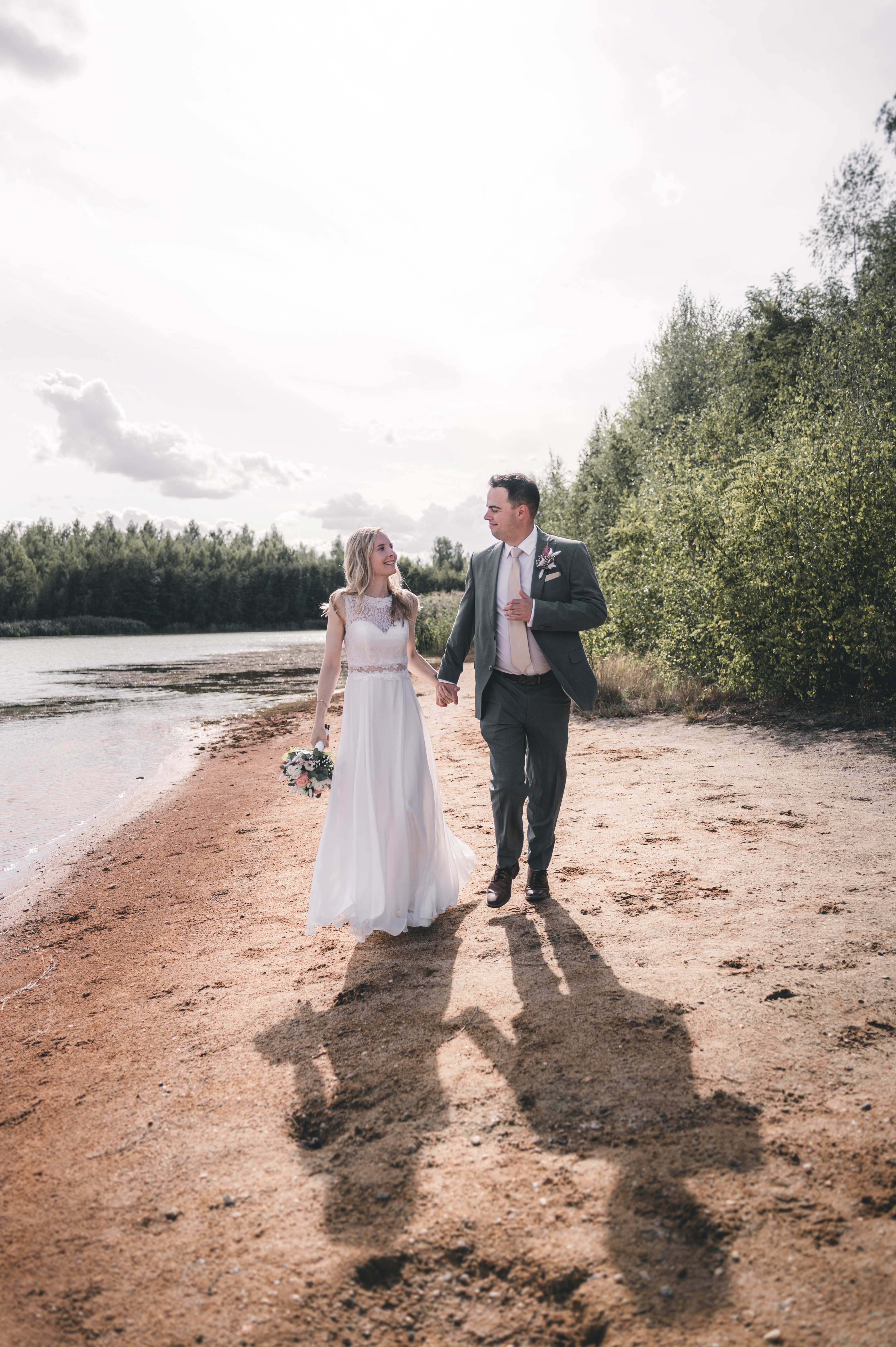 Das frischgebackene Ehepaar läuft Hand in Hand und glücklich lächelnd über den Sandstrand am Störmthaler See.