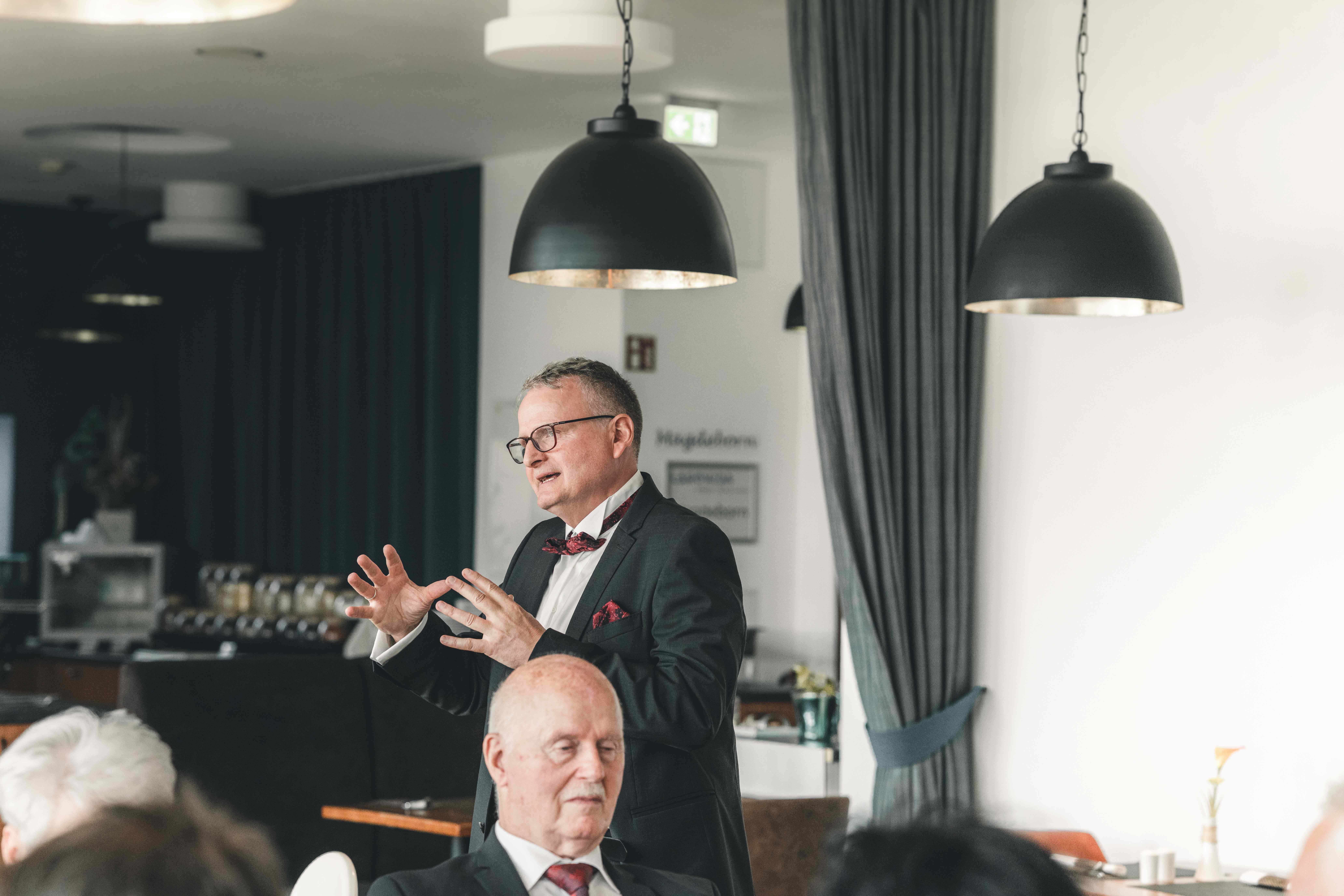 Der Brautvater im eleganten Anzug mit Fliege spricht gestikulierend vor den Gästen im Saal des Hotels am Störmthaler See.